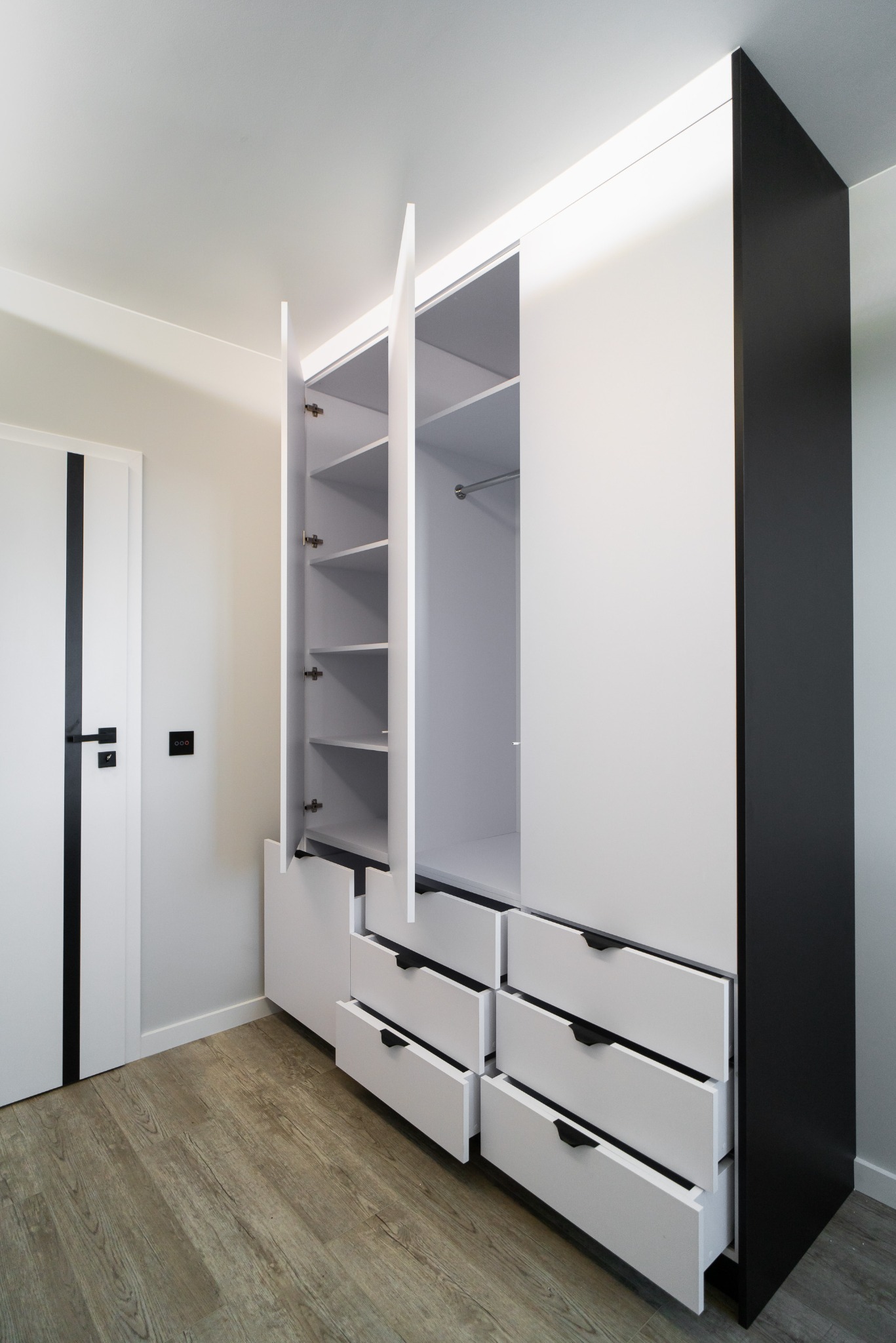 Nowoczesna szafa z białymi frontami i czarnym bokiem, częściowo otwarta, ukazująca półki i szuflady. Drzwi białe z czarnym paskiem i klamką w tle.