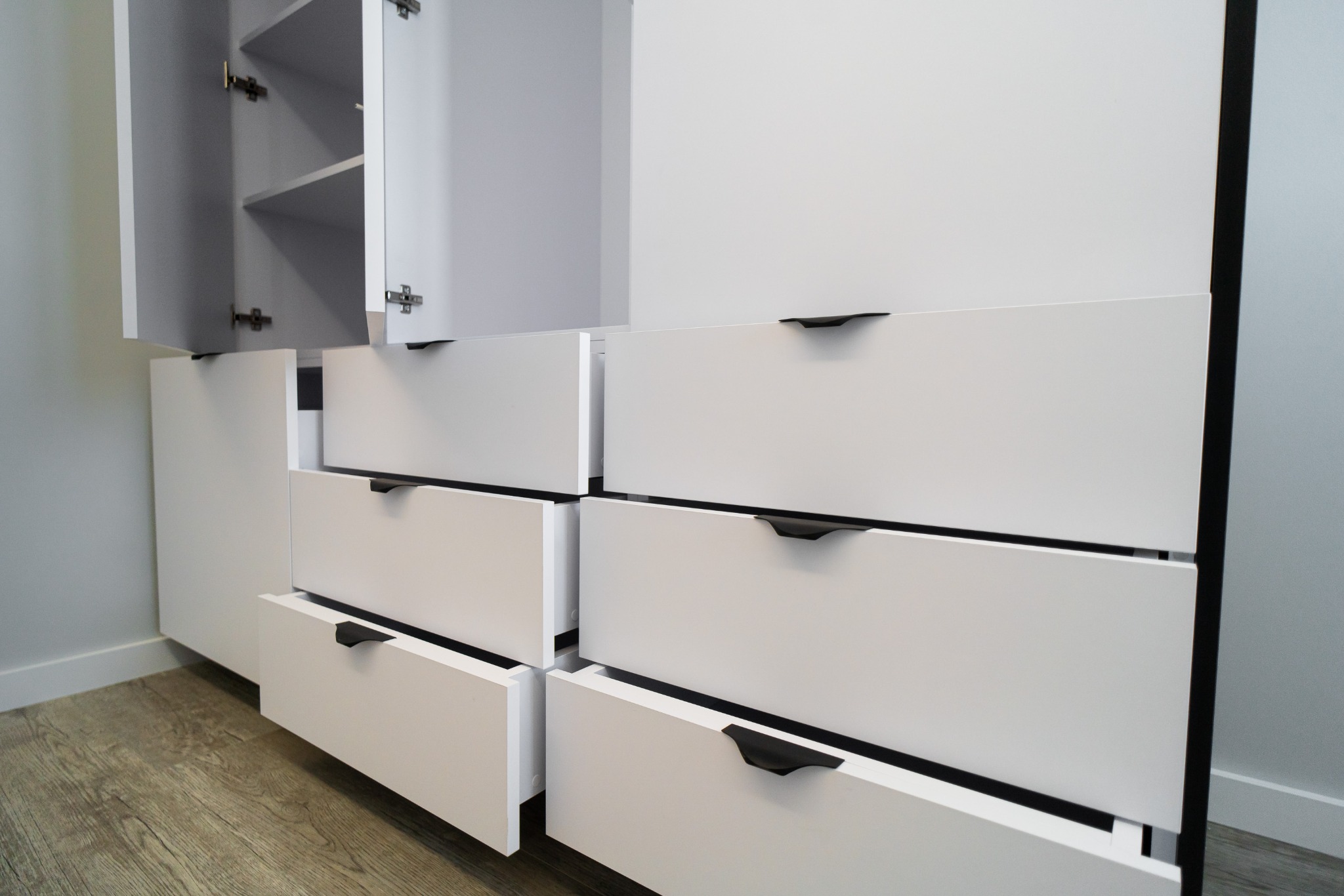 Biała komoda z szufladami i szafkami, czarne uchwyty, fronty otwarte. Widoczna podłoga z paneli. Minimalistyczny design, nowoczesne wykończenie.