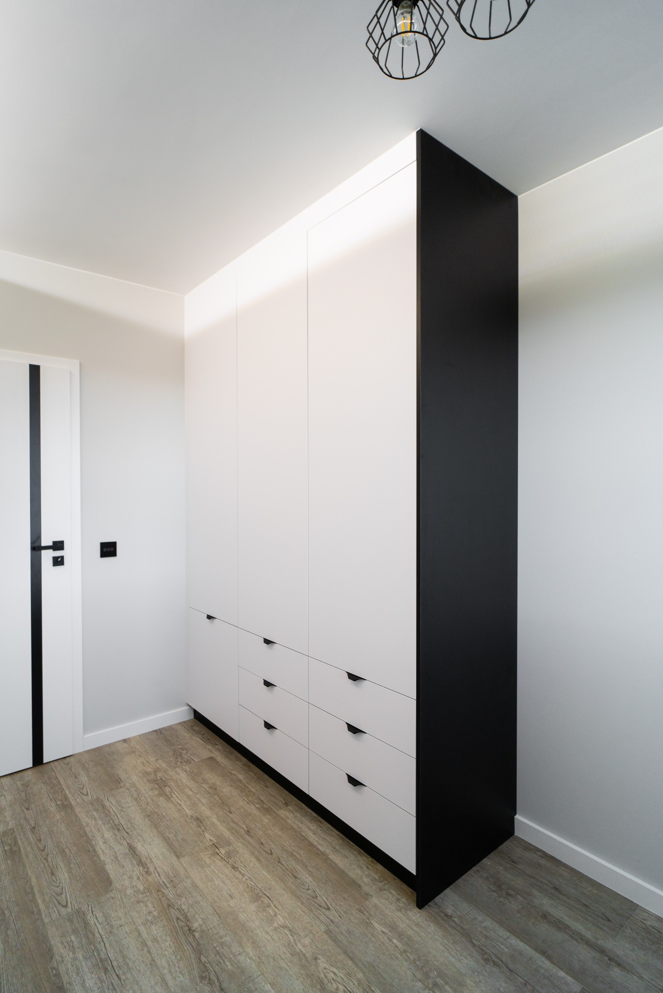 Biało-czarna szafa z szufladami i frontami bez uchwytów w nowoczesnym wnętrzu. Widoczna podłoga imitująca drewno i lampa sufitowa w stylu industrialnym.