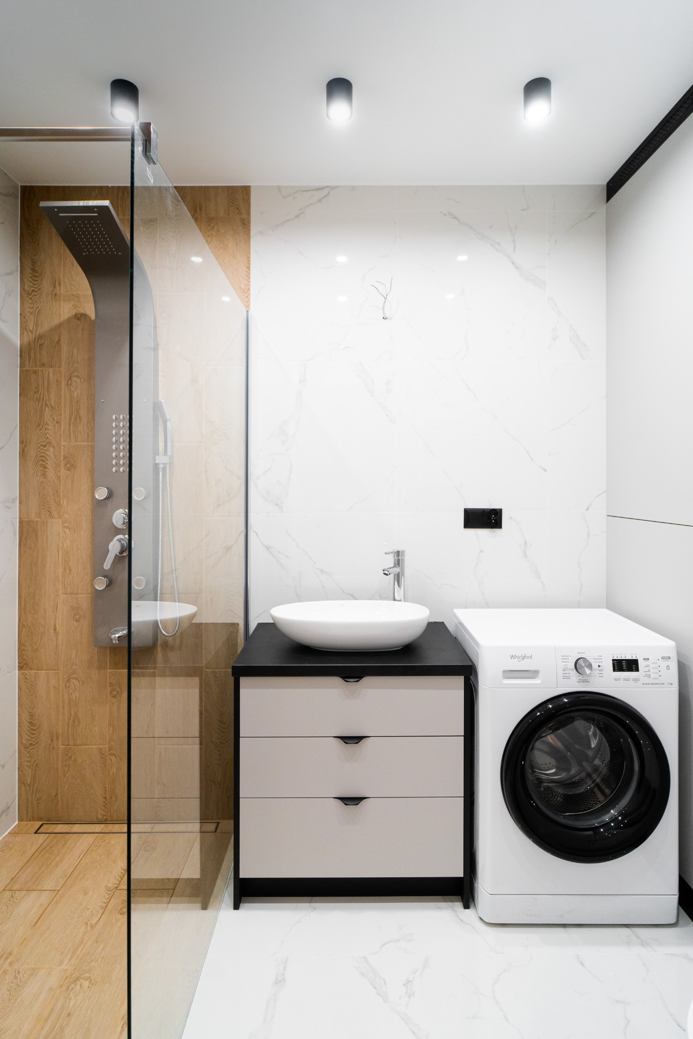 Nowoczesna łazienka z kabiną prysznicową typu walk-in, umywalką nablatową na szafce z szufladami i pralką. Białe marmurowe płytki i drewniane akcenty.