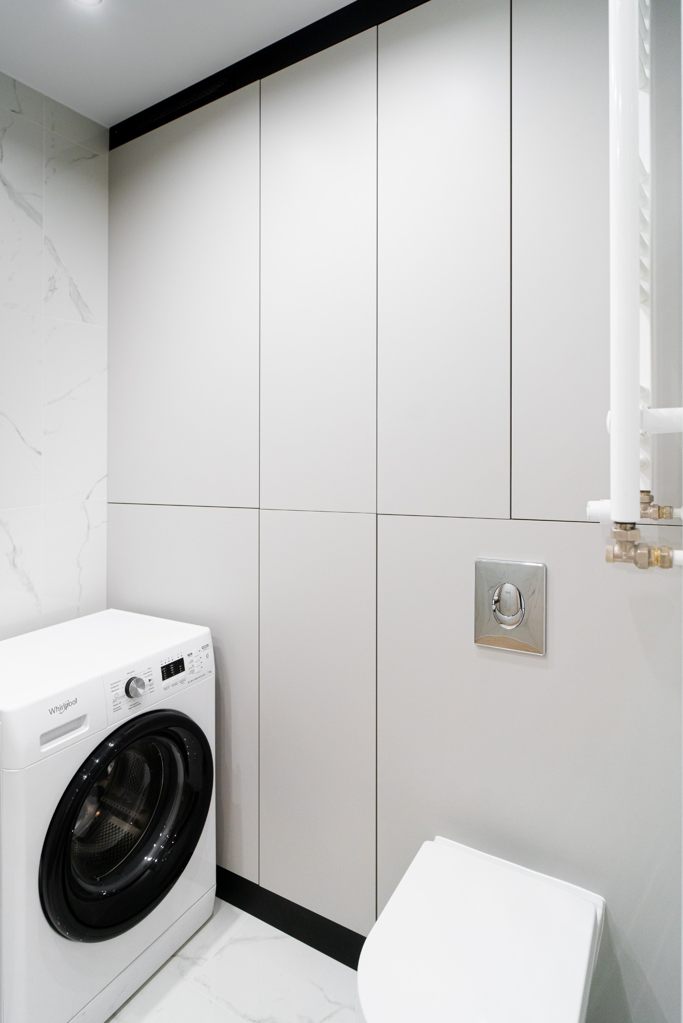Nowoczesna łazienka w bieli i szarości z pralką Whirlpool, zabudową meblową z czarnymi akcentami i marmurową posadzką. Minimalistyczny design, eleganckie wykończenie.