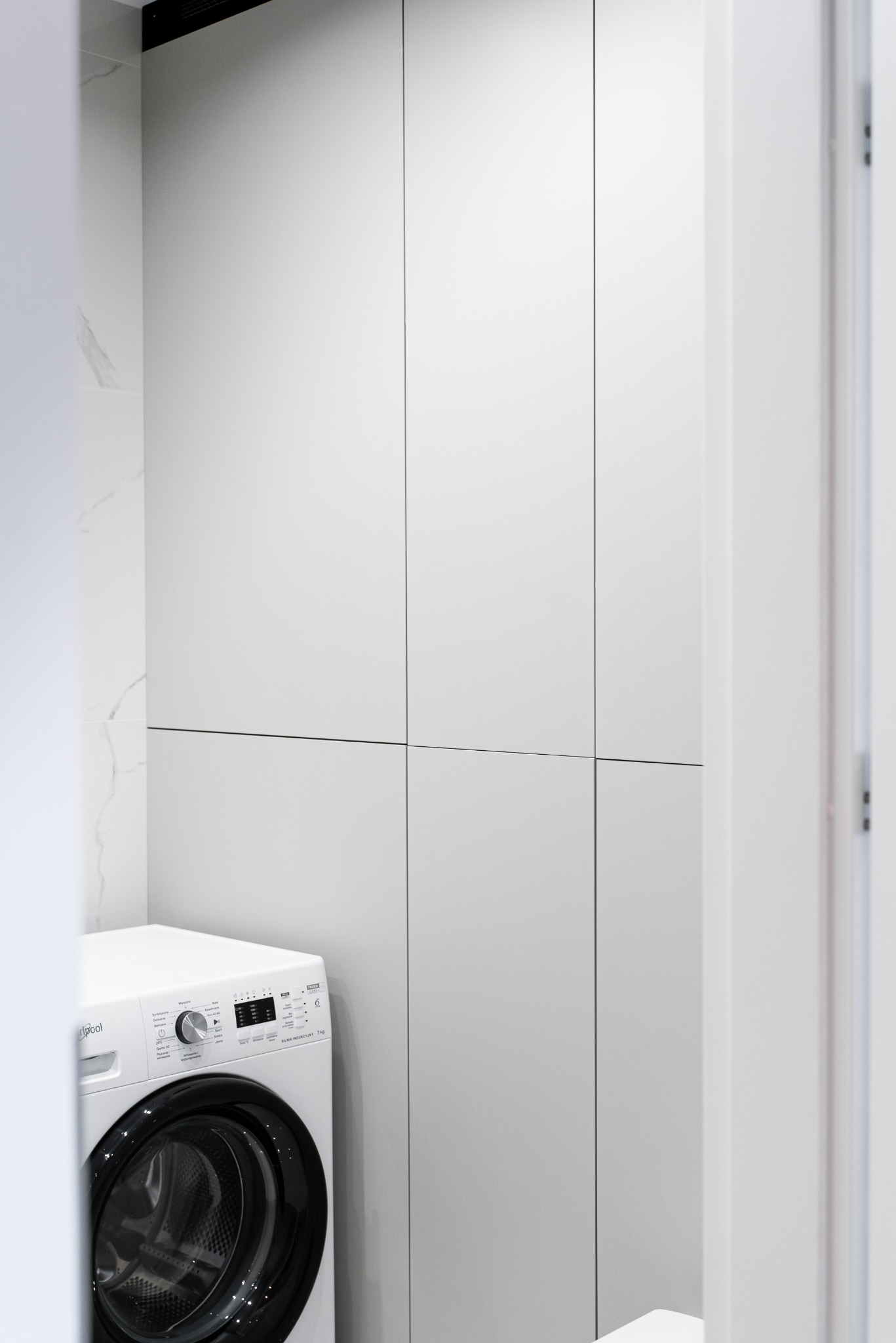 Minimalistyczna łazienka: biała pralka Whirlpool obok zabudowy szafy z gładkimi, jasnymi frontami, marmurowe płytki. Nowoczesne wnętrze.