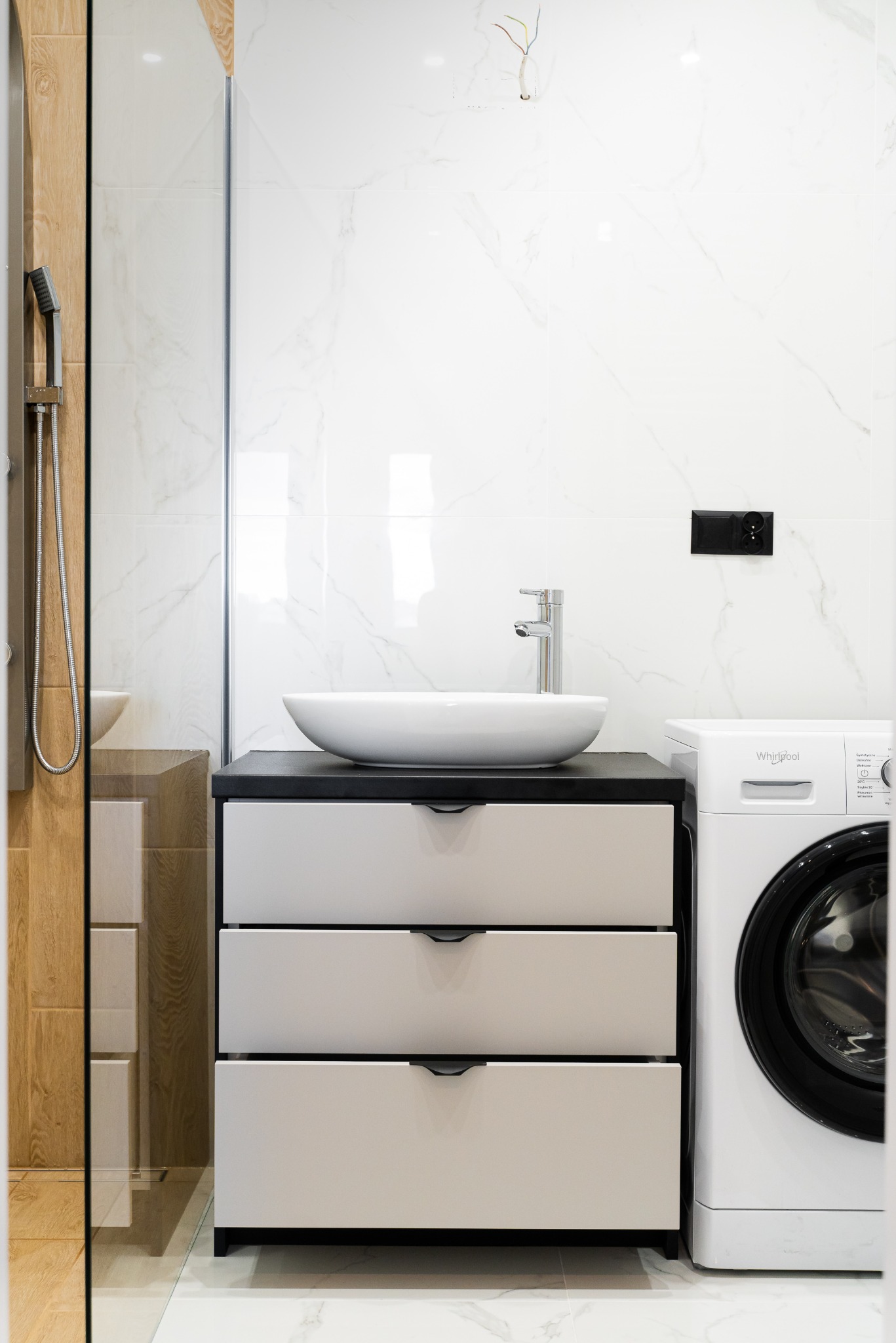 Nowoczesna łazienka z umywalką nablatową na szafce z szufladami, obok pralka. Białe płytki imitujące marmur, szklana kabina prysznicowa z drewnianymi akcentami.