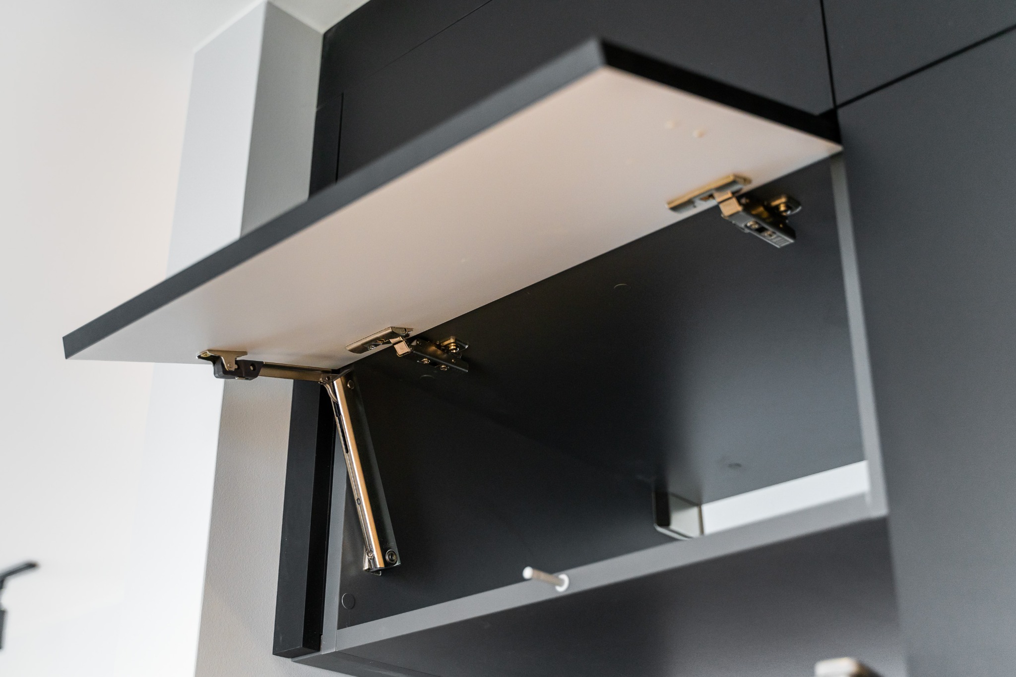 Otwarta szafka kuchenna z mechanizmem podnoszenia, front biały z czarnym obramowaniem, widoczne zawiasy i siłownik. Minimalistyczny design.