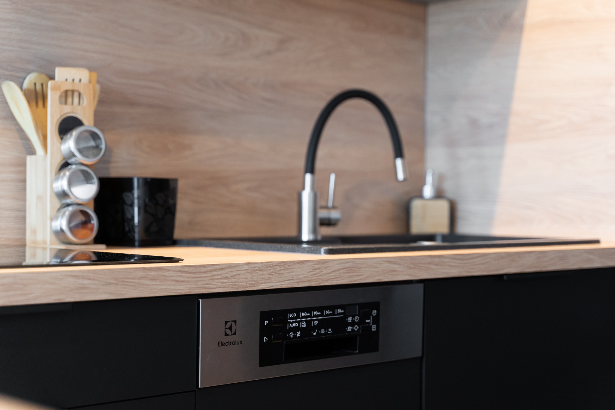 Minimalistyczna kuchnia: czarne szafki, drewniany blat, metalowa bateria i akcesoria. Widoczny panel sterowania zmywarki Electrolux.