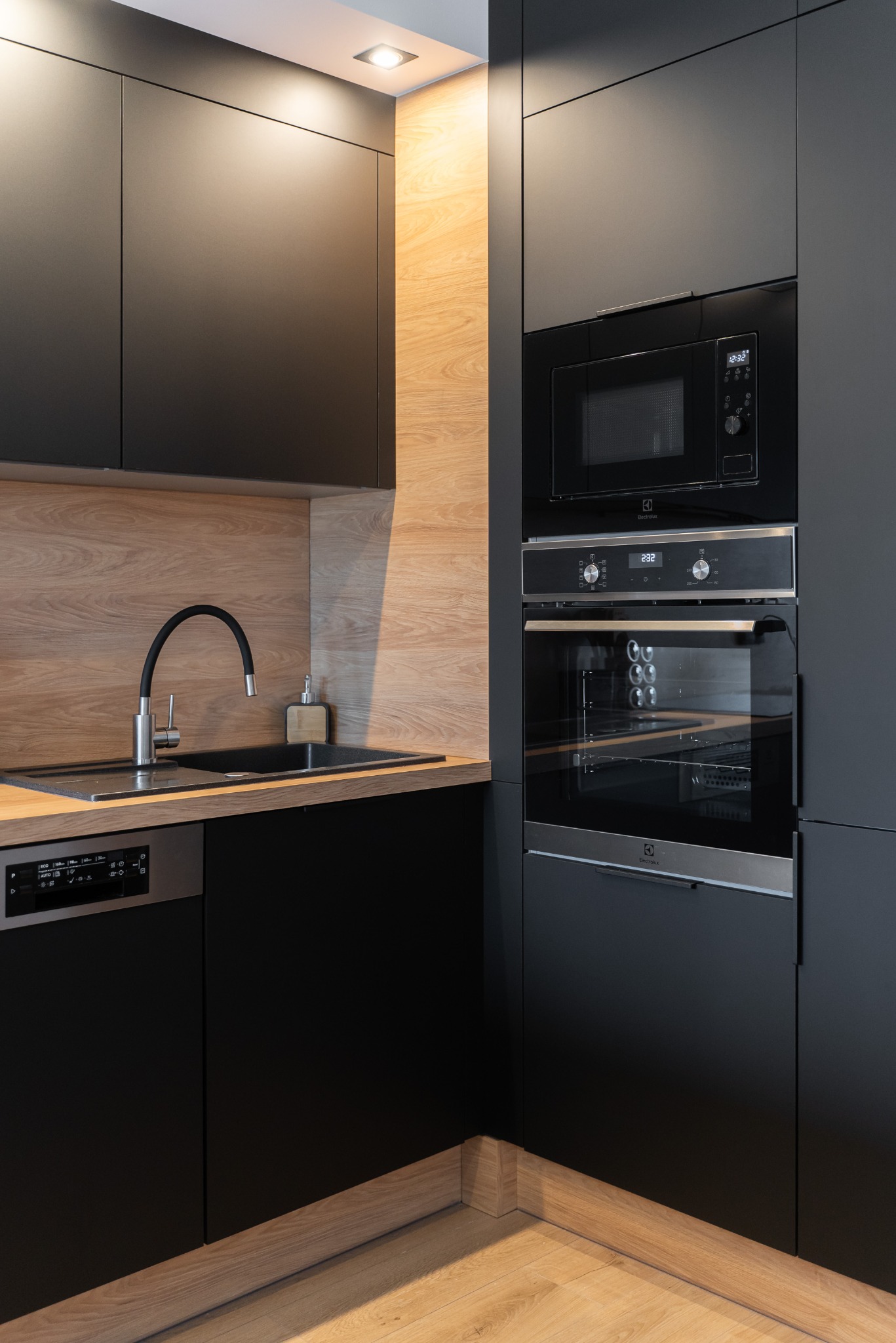 Nowoczesna, czarna kuchnia z drewnianymi akcentami: zabudowa z piekarnikiem i mikrofalówką, czarny zlew z baterią, drewniany blat i podłoga. Minimalistyczny design.