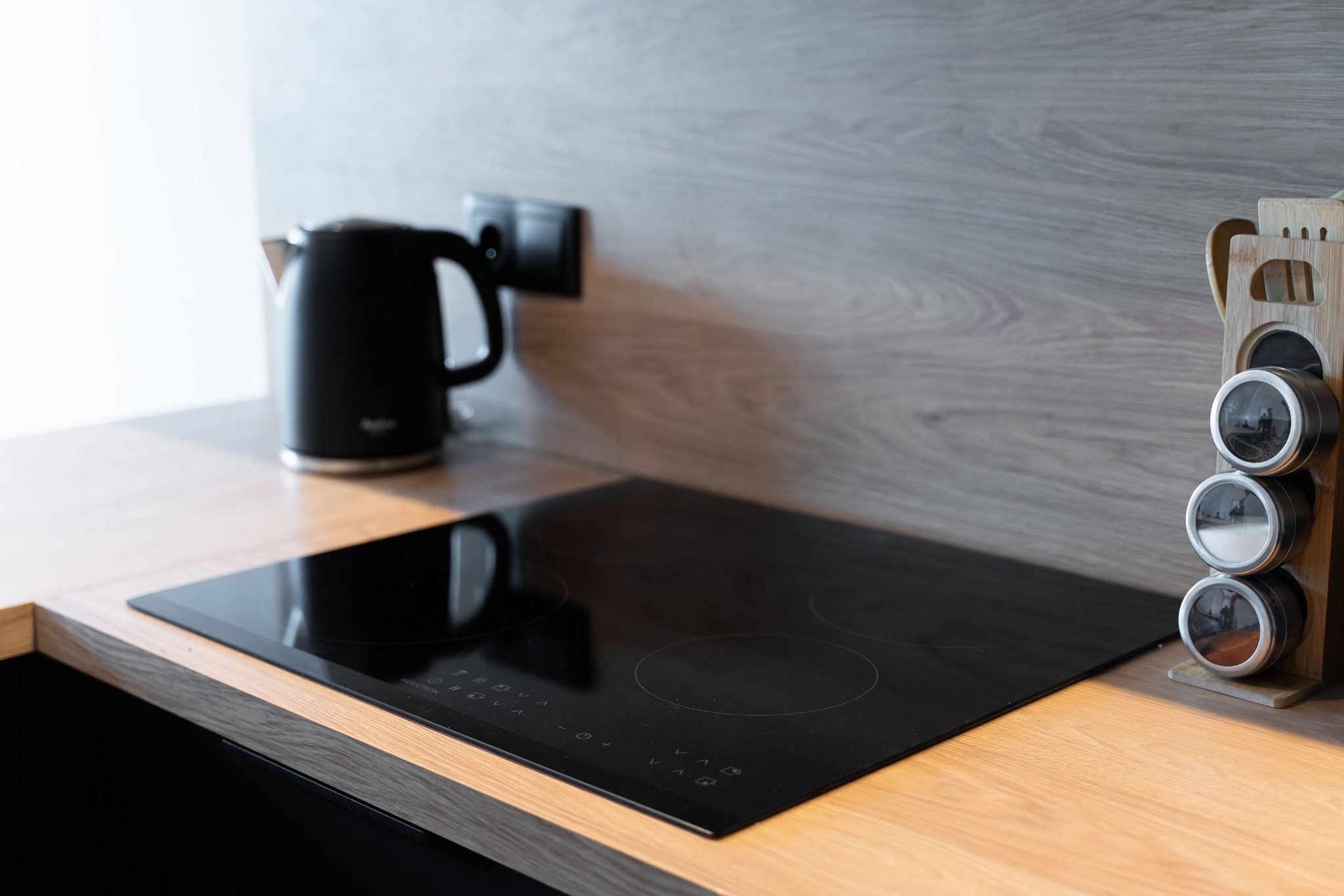 Czysta, nowoczesna kuchnia: czajnik, płyta indukcyjna i przyprawnik na blacie z drewnianym wykończeniem i szarą ścianą. Minimalistyczny design.