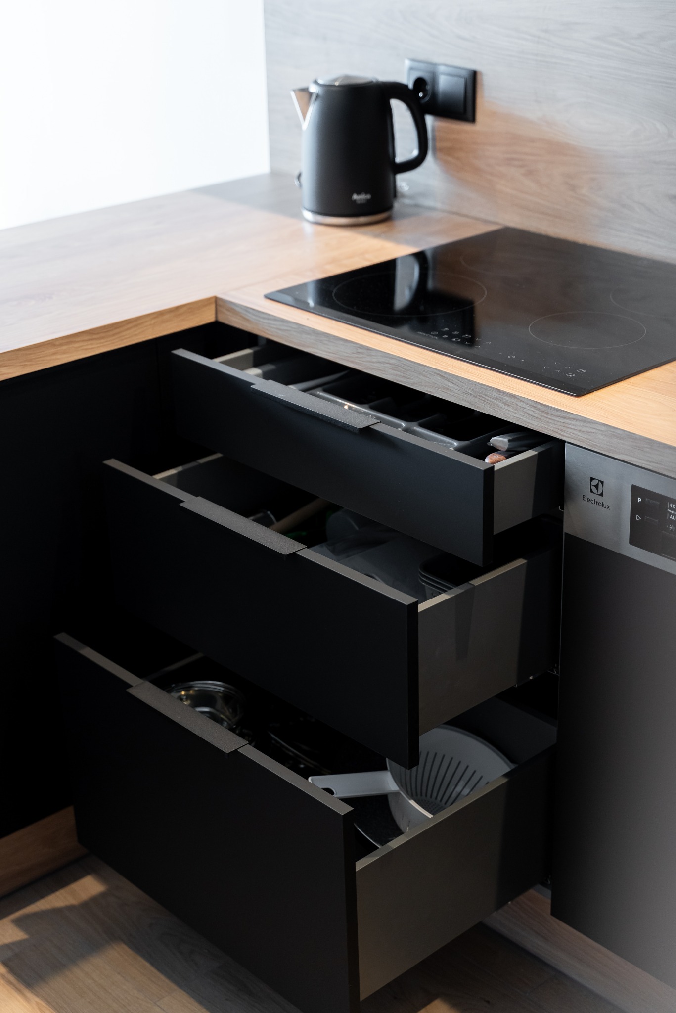 Ciemna kuchnia z otwartymi szufladami, w których widać naczynia. Na blacie czajnik i płyta indukcyjna. Minimalistyczny design, drewniany blat, nowoczesne wykończenie.