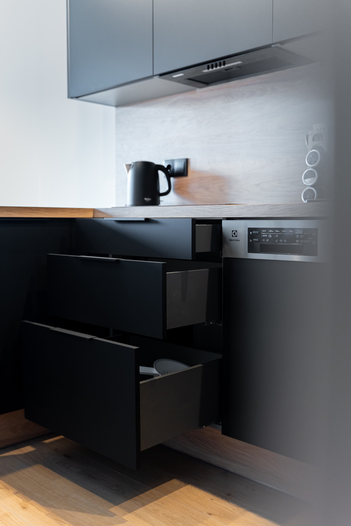 Minimalistyczna kuchnia: czarne, matowe fronty szuflad z otwartą dolną szufladą, drewniany blat, czajnik i srebrna zmywarka Electrolux. Nowoczesny design.