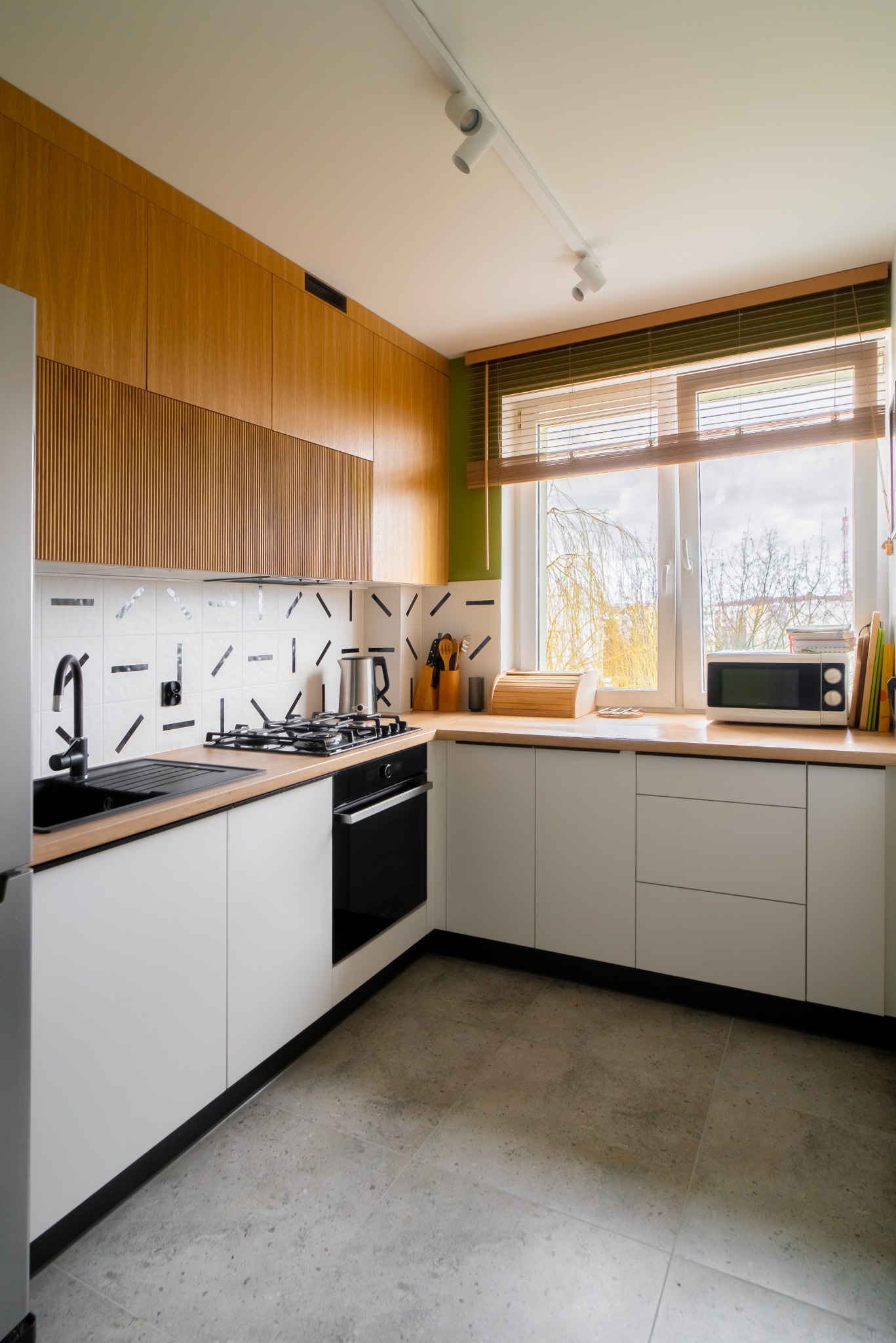 Nowoczesna kuchnia: białe szafki dolne, drewniane górne z ryflowanym panelem, czarny zlew i płyta, geometryczne płytki, okno z żaluzjami. Jasne wnętrze.