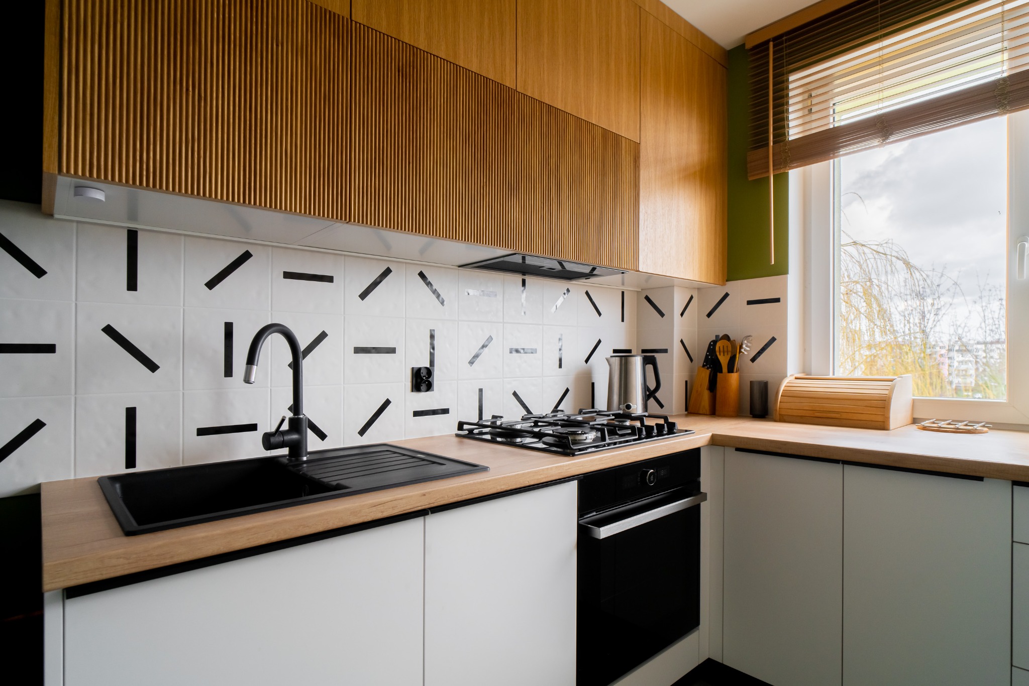 Nowoczesna kuchnia z drewnianymi szafkami górnymi, białymi dolnymi, blatem z drewna i czarną armaturą na tle białych płytek z czarnymi wzorami.