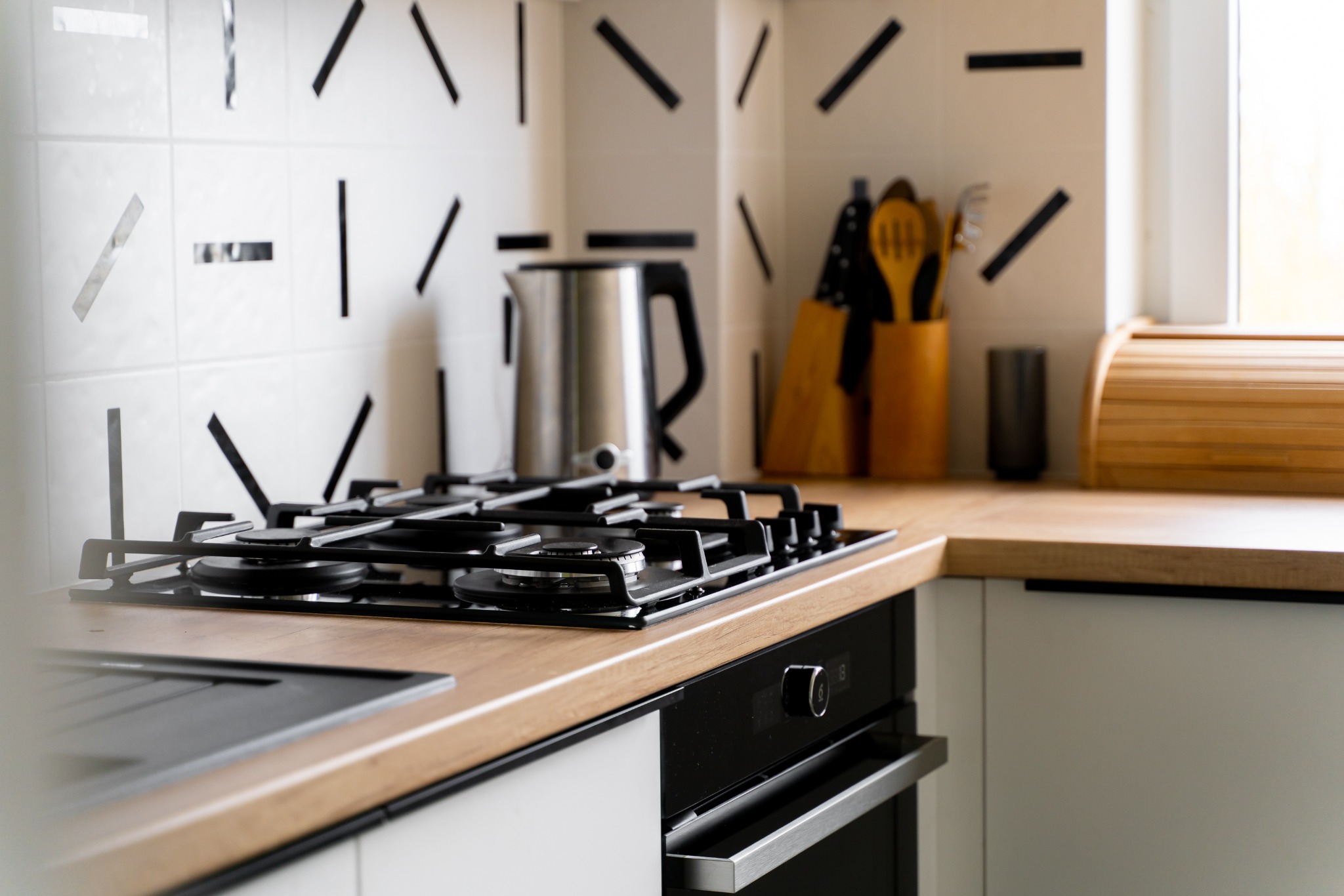 Nowoczesna kuchnia z blatem drewnopodobnym i płytą gazową w Gdańsku. Białe szafki, oryginalne płytki na ścianie, czajnik i akcesoria kuchenne.