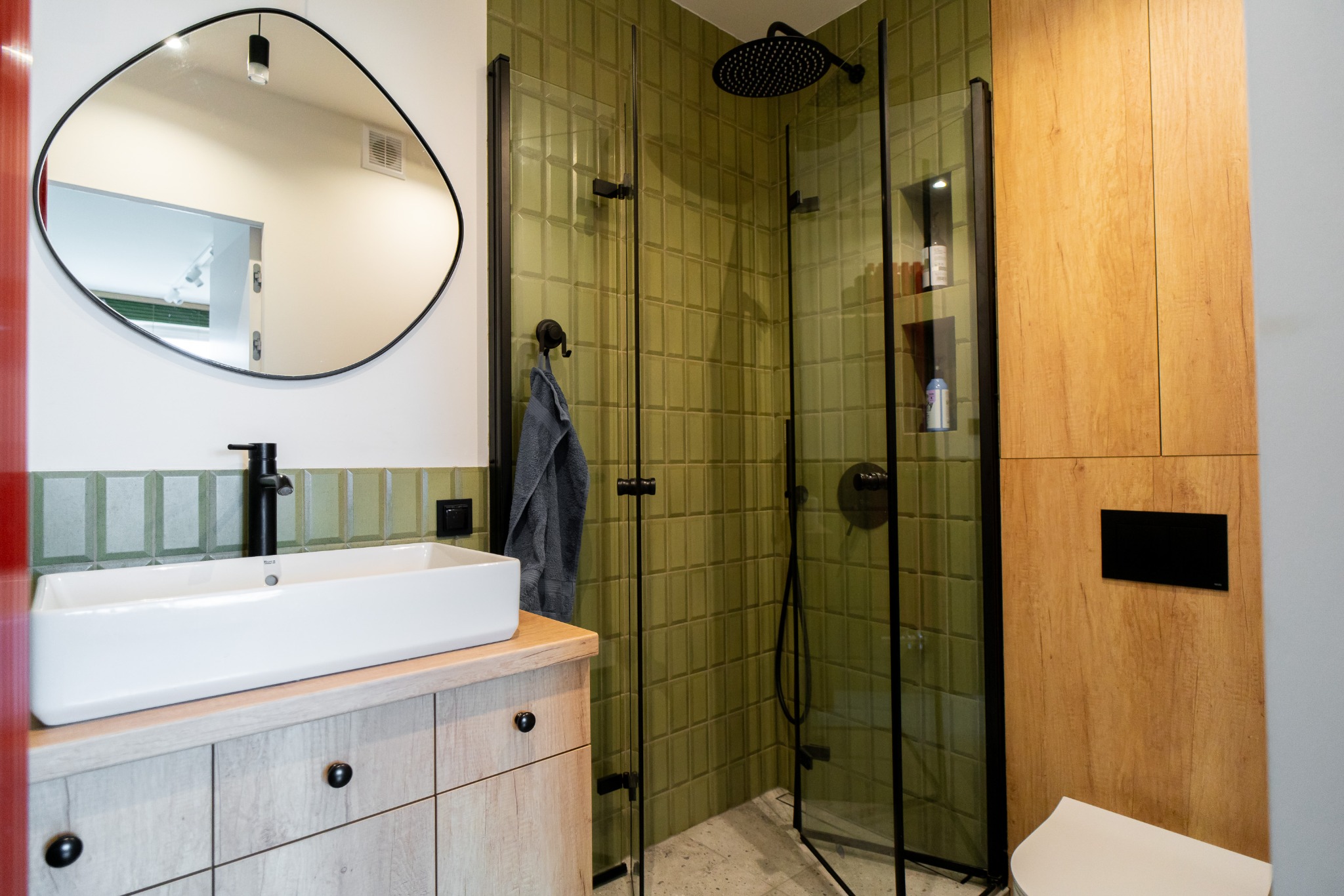 Nowoczesna łazienka z zielonymi płytkami, kabiną prysznicową typu walk-in, umywalką nablatową na szafce z drewnianymi frontami i asymetrycznym lustrem.