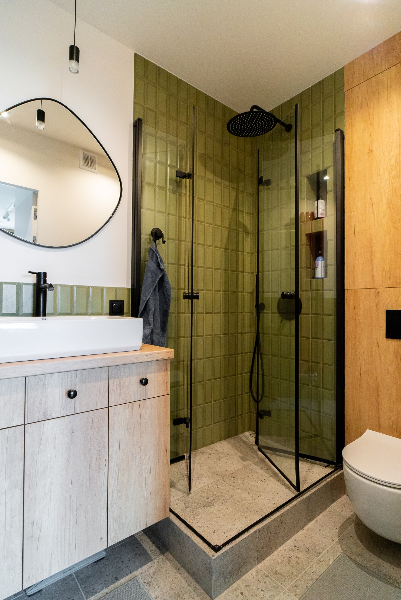 Nowoczesna łazienka z kabiną prysznicową typu walk-in, umywalką na szafce z drewnopodobnym frontem i zielonymi płytkami na ścianie. Czarna armatura i rama kabiny.