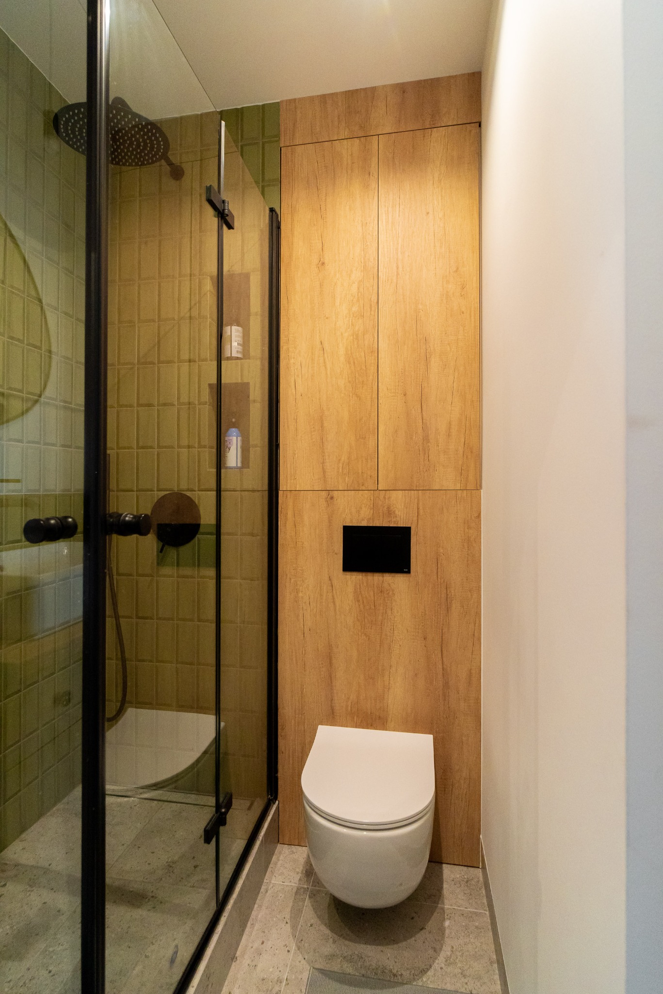 Nowoczesna łazienka z kabiną prysznicową typu walk-in, toaletą podwieszaną i drewnianą zabudową. Użyto płytek w odcieniach zieleni i szarości.