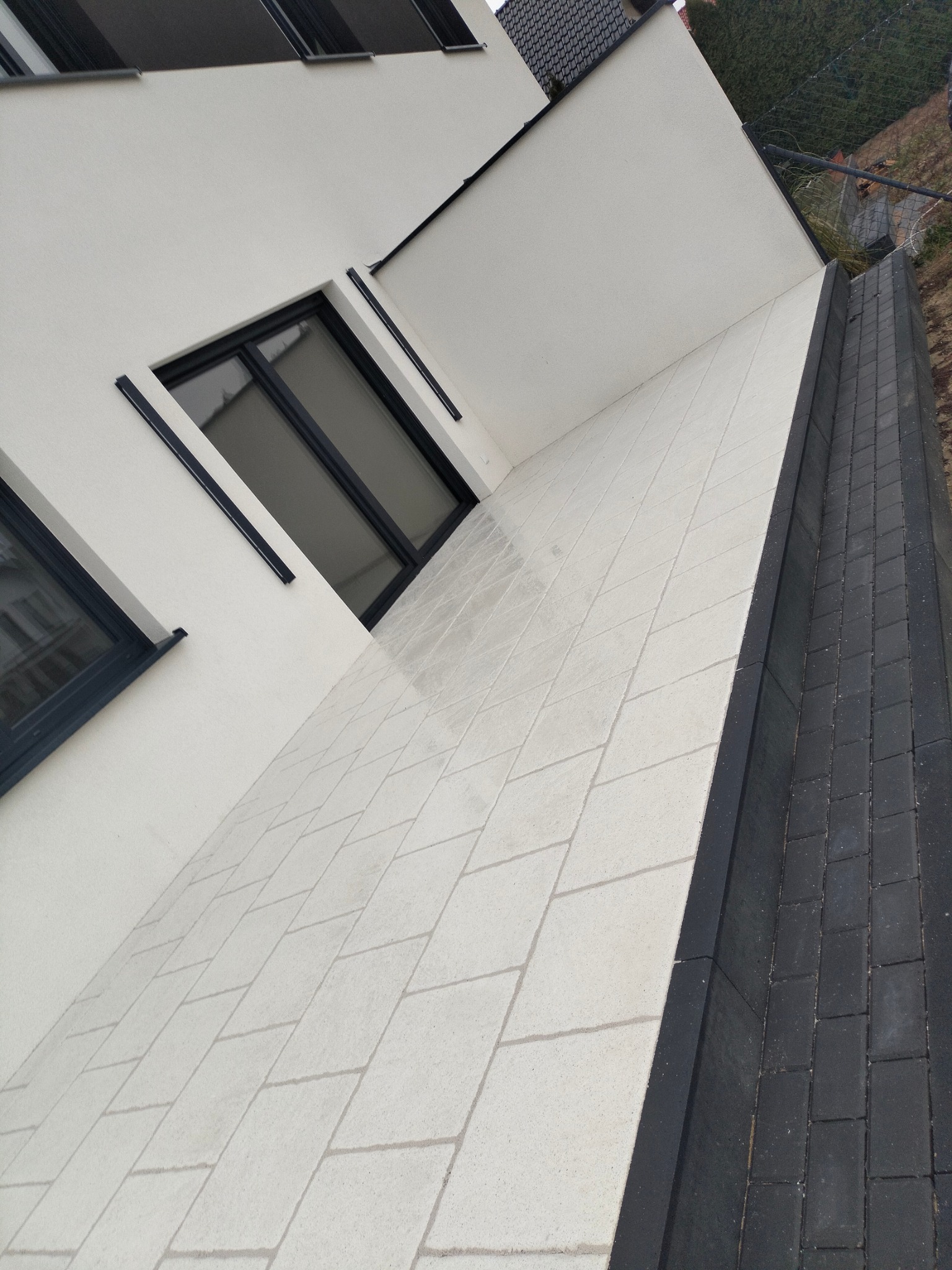 Nowoczesny chodnik z jasnej kostki brukowej ułożonej przy białej ścianie budynku z czarnymi ramami okiennymi i ciemnymi obrzeżami.