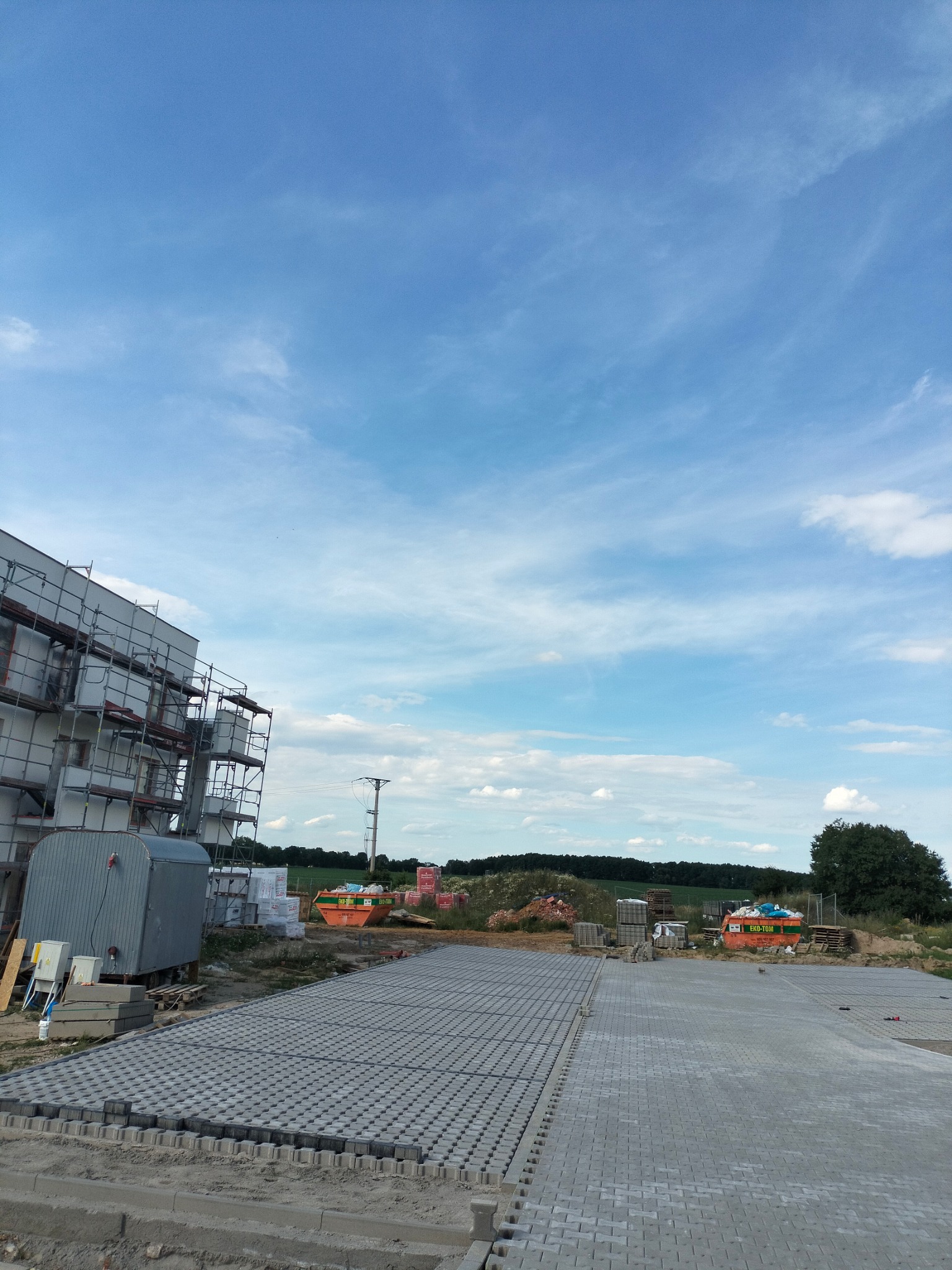 Powierzchnia wyłożona szarą kostką brukową w trakcie prac, widoczne przygotowane materiały budowlane, rusztowania przy budynku w tle i błękitne niebo.