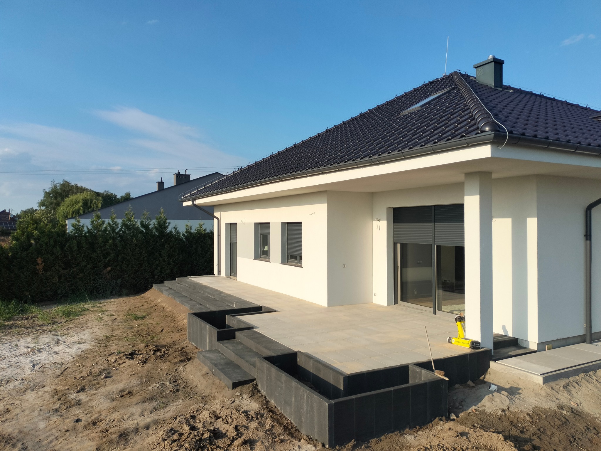 Jasne płytki tarasowe ułożone przed nowoczesnym domem z ciemną dachówką, widoczne schody z kostki i narzędzia brukarskie.
