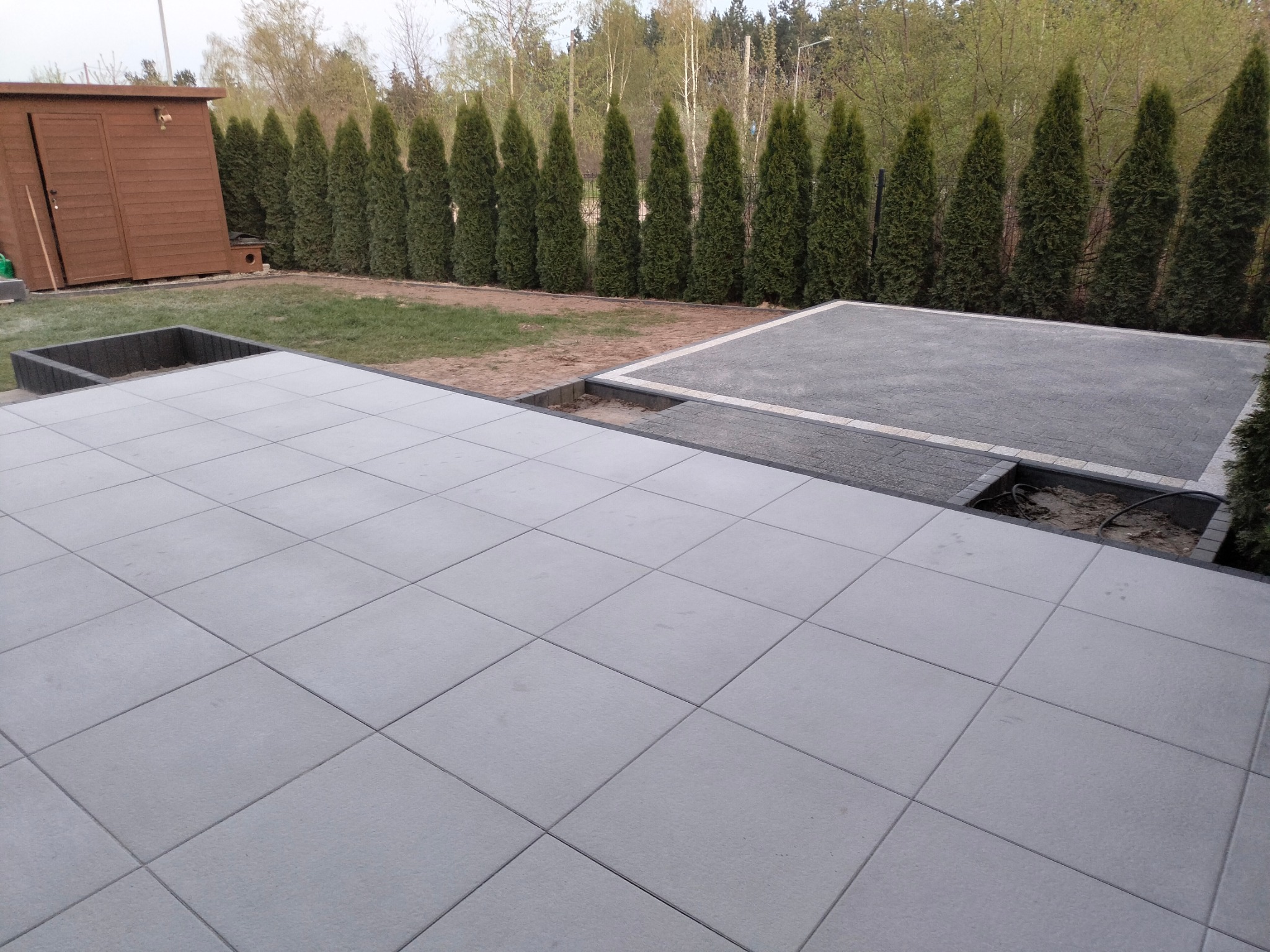 Ułożona nawierzchnia z jasnoszarych, kwadratowych płyt betonowych, częściowo uzupełniona ciemniejszą kostką, widoczny fragment rabaty i rząd tuj w tle.