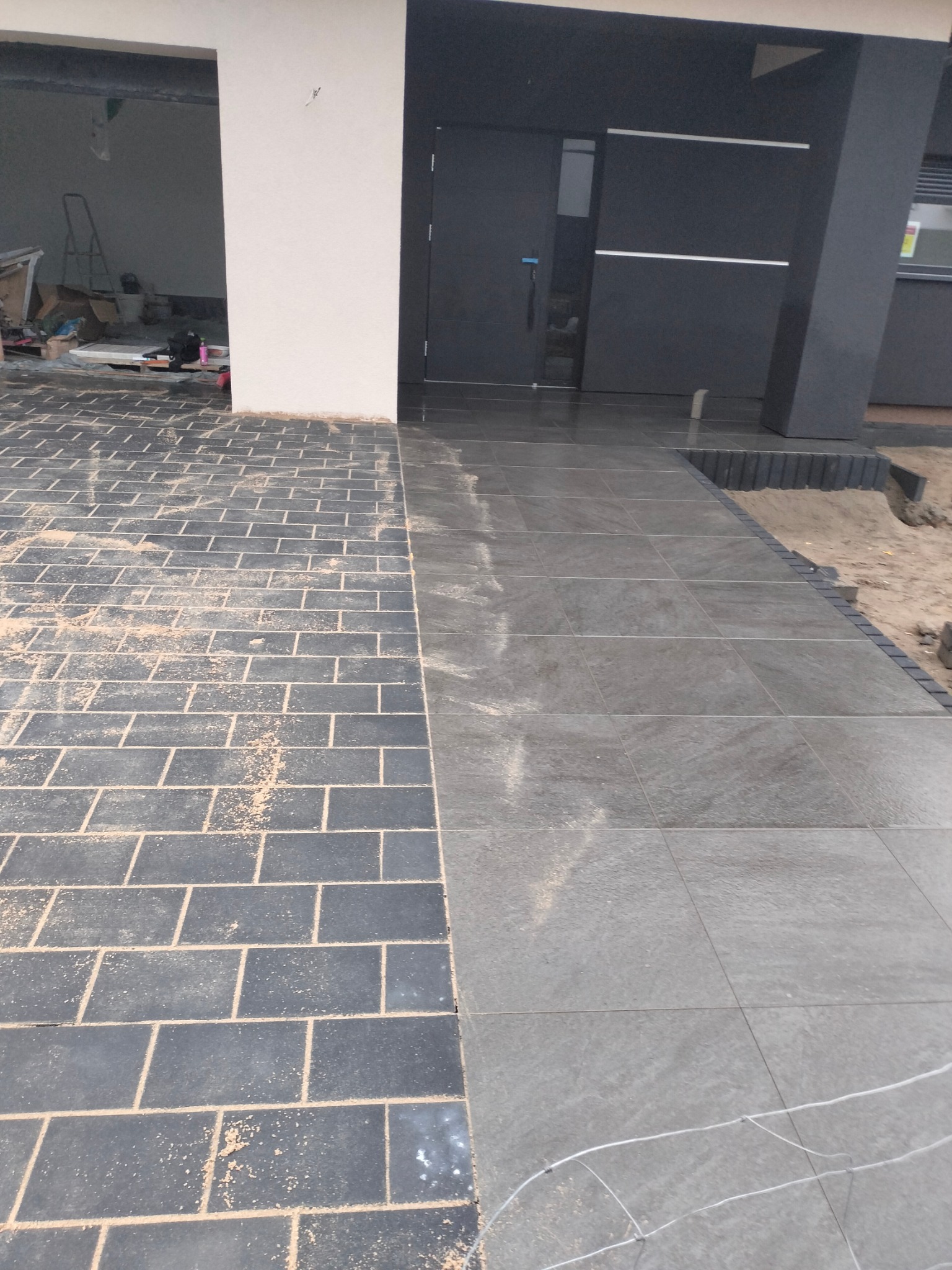 Nowo ułożona, częściowo zasypana piaskiem, ciemnoszara kostka brukowa w kształcie cegiełek, sąsiadująca z płytami chodnikowymi w odcieniach szarości prowadzącymi do nowoczesnego domu z ciemnymi...