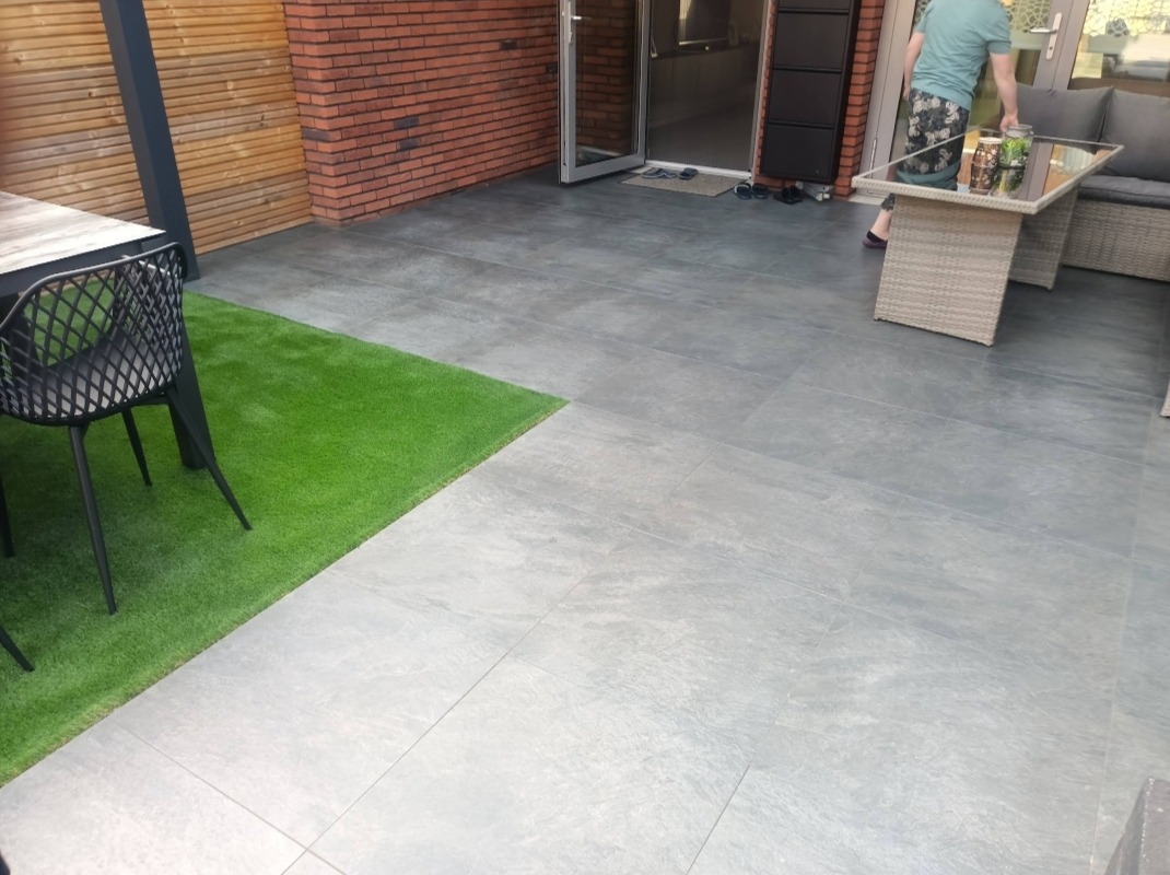 Szara, duża płytka na tarasie z fragmentem sztucznej trawy, stolikiem kawowym i sofą, widok na drzwi tarasowe i fragment drewnianego ogrodzenia.