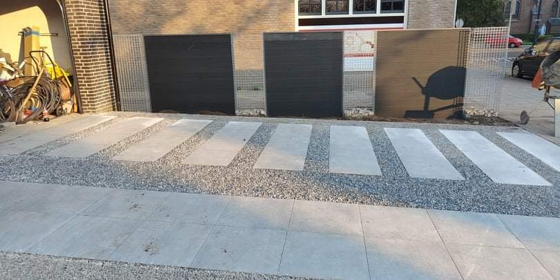 Nowoczesna ścieżka z szarych płyt betonowych i drobnego żwiru prowadząca do budynku z ciemnymi panelami elewacyjnymi i koszem gabionowym. Widoczne narzędzia i sprzęt budowlany po lewej stronie.