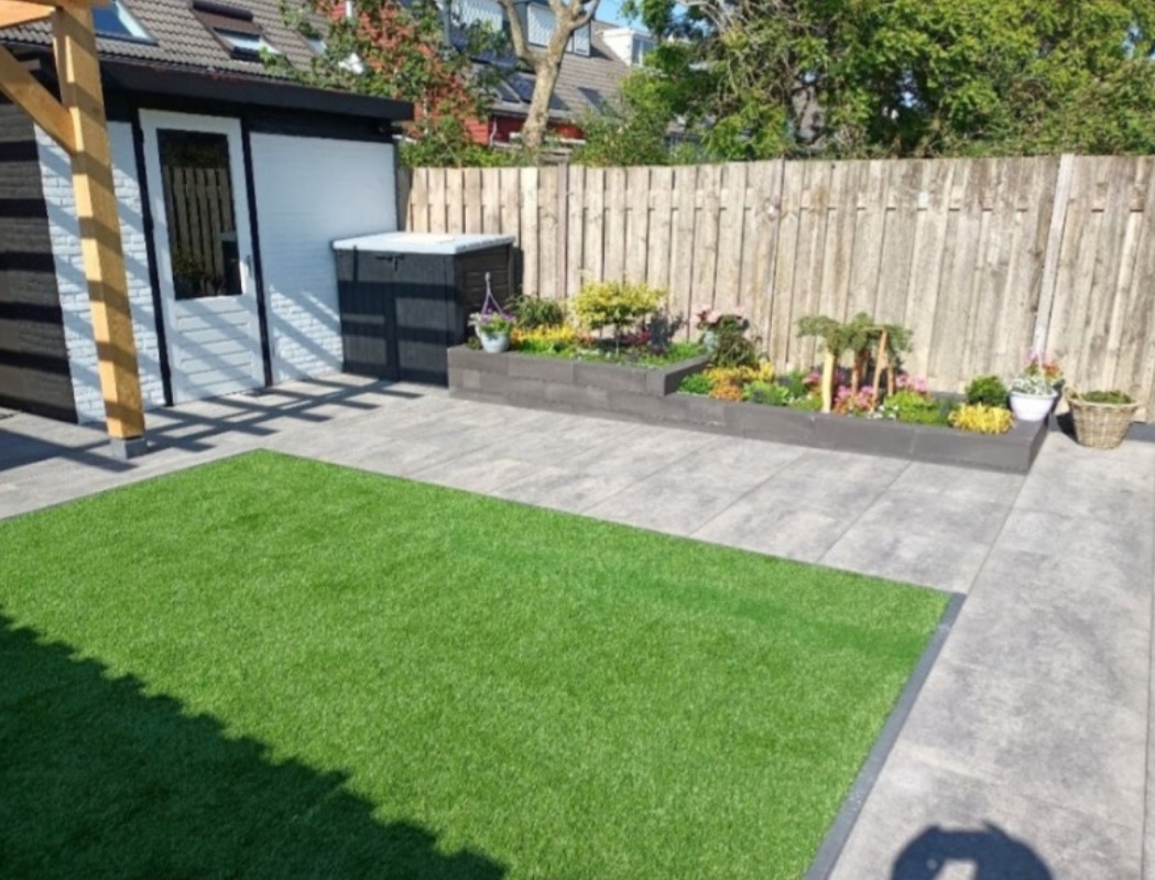 Ułożona szara kostka brukowa tworząca patio z fragmentem sztucznej trawy, drewniany domek narzędziowy z białymi drzwiami, podwyższona rabata kwiatowa z roślinami i drewniany płot w tle.