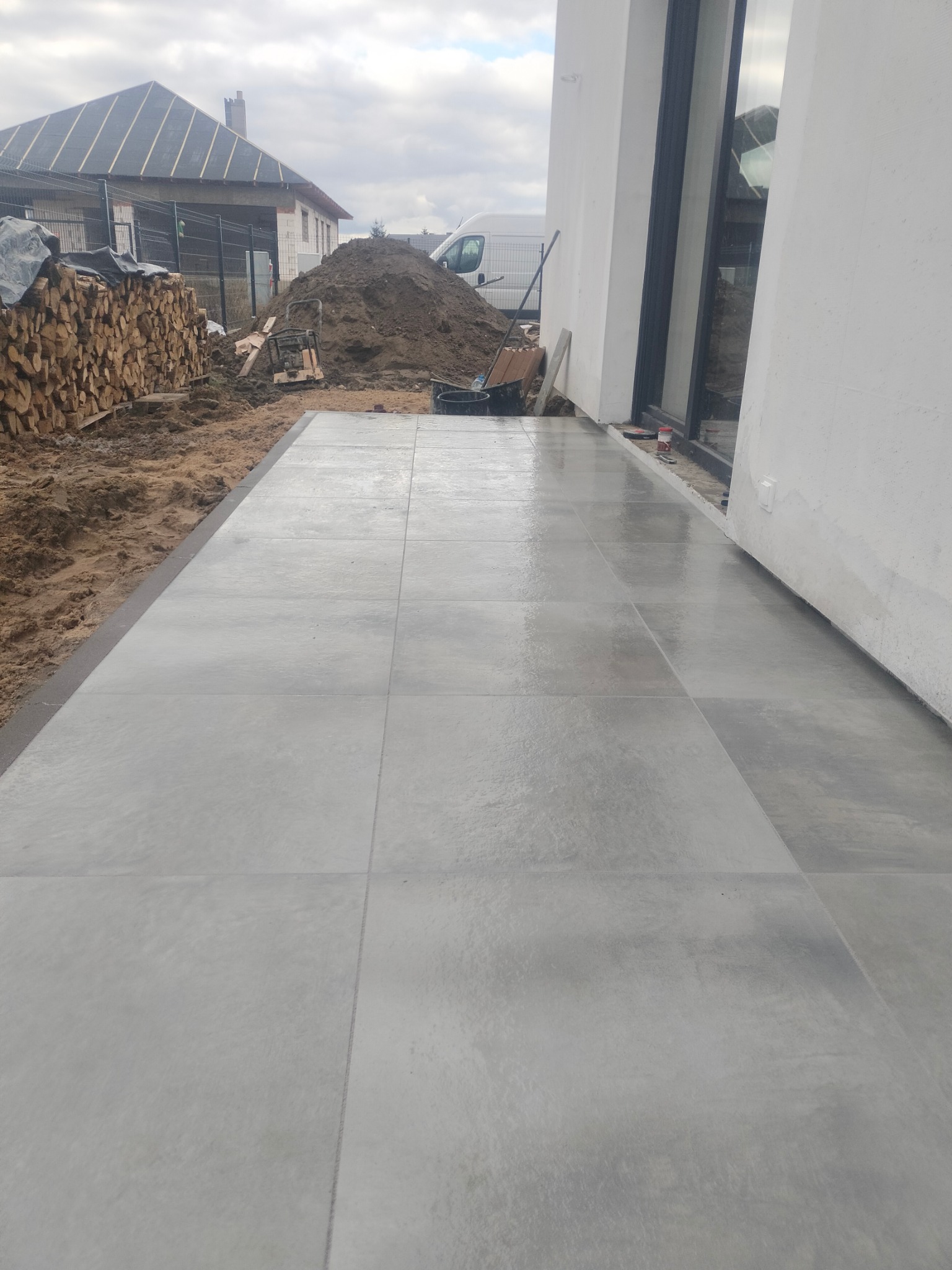 Świeżo ułożona, duża, szara kostka brukowa na tarasie w Murowanej Goślinie, widoczna sterta ziemi i przygotowane drewno opałowe w tle.