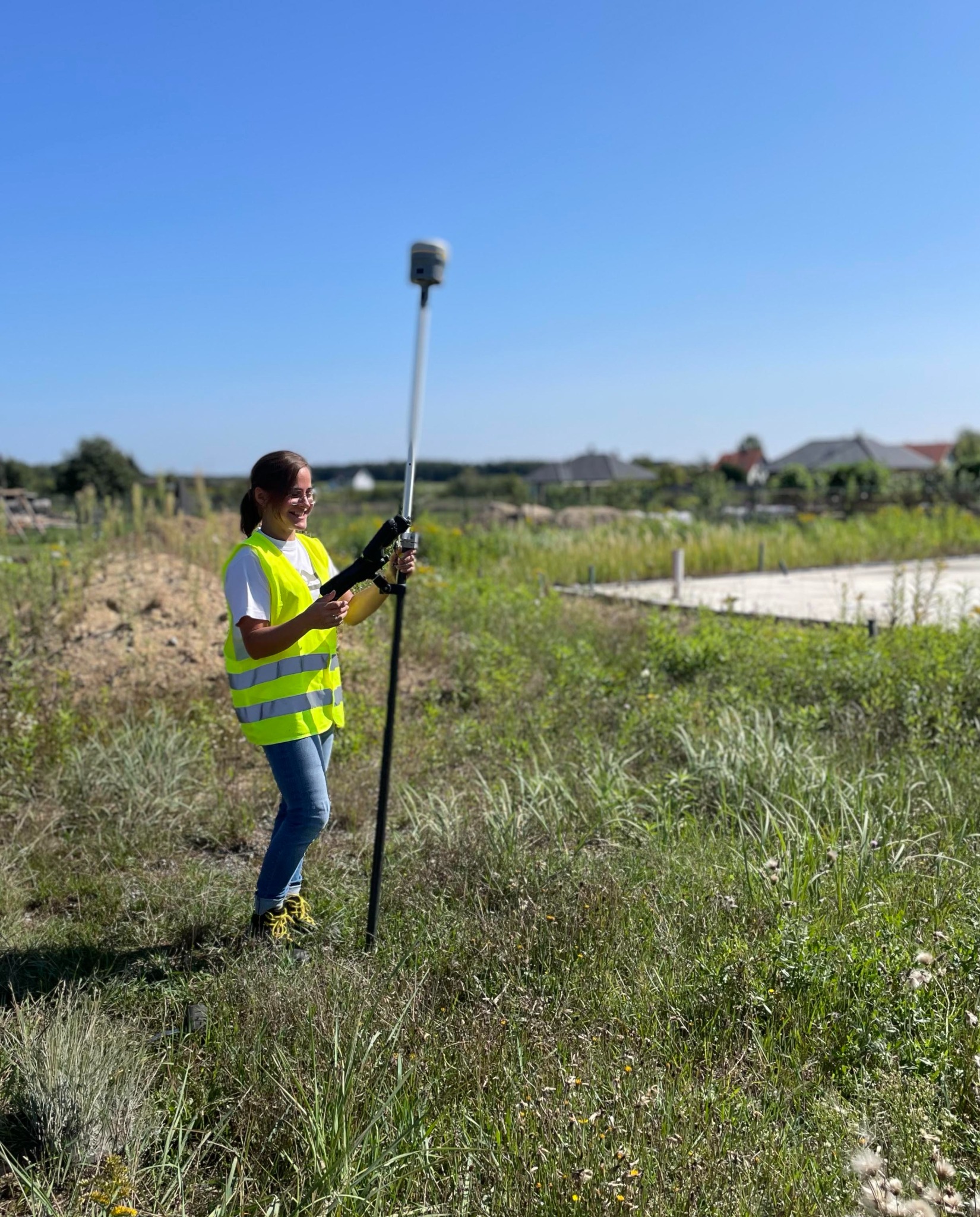 Kobieta w kamizelce odblaskowej obsługuje precyzyjny instrument pomiarowy na tyczce, w tle widoczne fundamenty budynków i błękitne niebo.