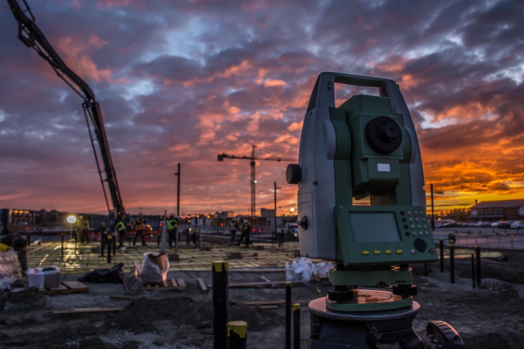 Teodolit na statywie na placu budowy o zmierzchu, w tle widoczna pompa do betonu i pracownicy budowlani, niebo z pomarańczowymi chmurami.