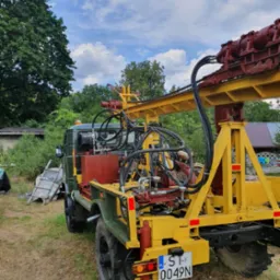 Żółto-czerwona wiertnica zamontowana na zielonym pojeździe terenowym, z tablicą rejestracyjną ST 0049N, na trawiastym terenie z drzewami w tle i błękitnym niebem.