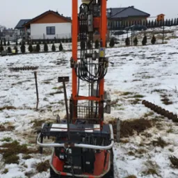 Wiertnica do studni na śniegu, widoczne ślady opon i elementy wiertła, w tle zabudowa jednorodzinna