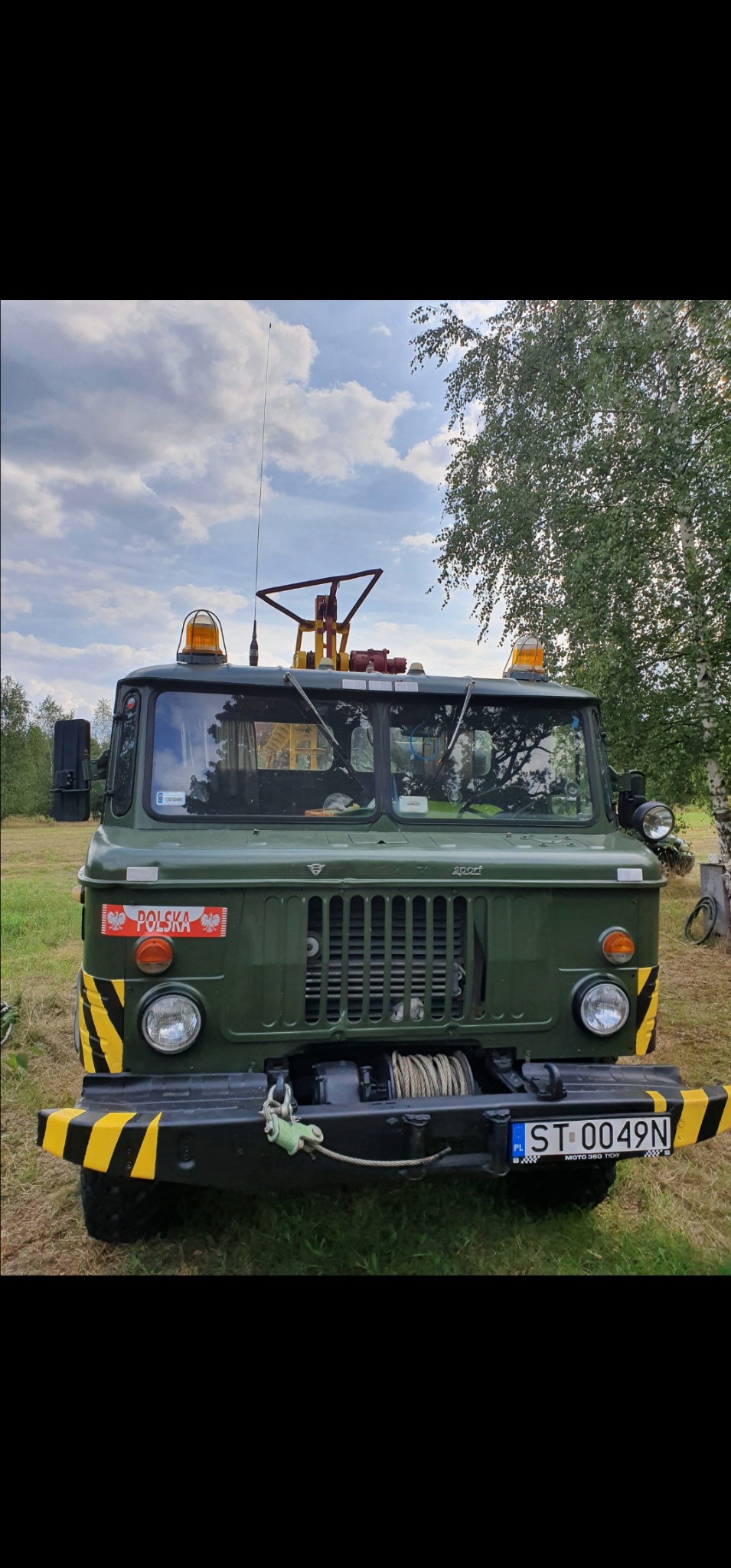 Zielony samochód ciężarowy z wyciągarką i żółto-czarnymi pasami ostrzegawczymi na zderzaku, zaparkowany na trawiastym terenie z drzewami w tle.
