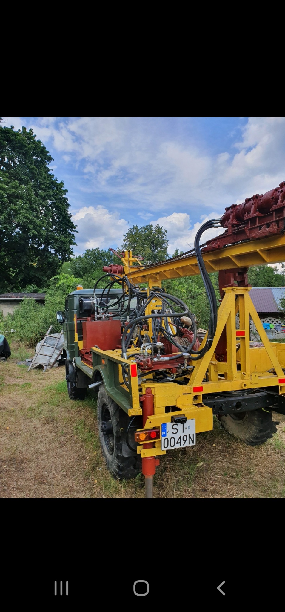 Żółto-czerwona wiertnica zamontowana na zielonym pojeździe terenowym, z tablicą rejestracyjną ST 0049N, na trawiastym terenie z drzewami w tle i błękitnym niebem.