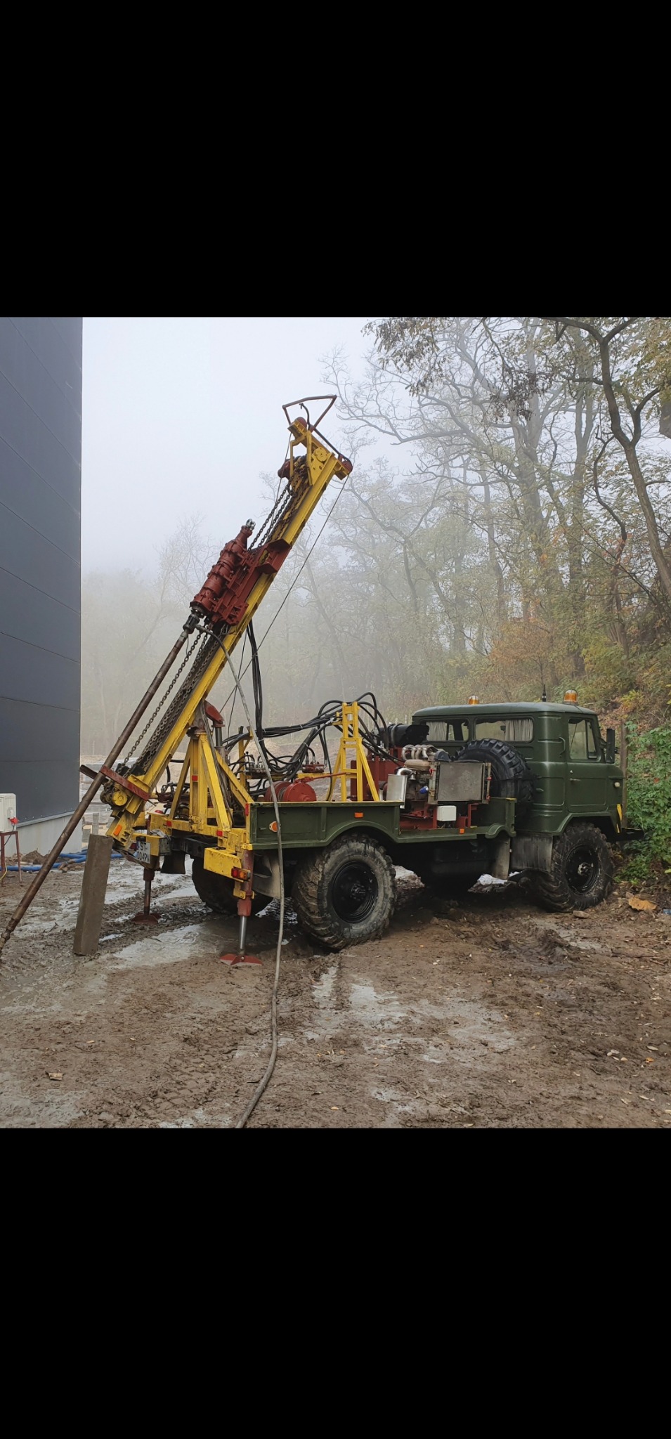 Zielony samochód ciężarowy z żółtą wiertnicą na błotnistym terenie, w tle drzewa we mgle.