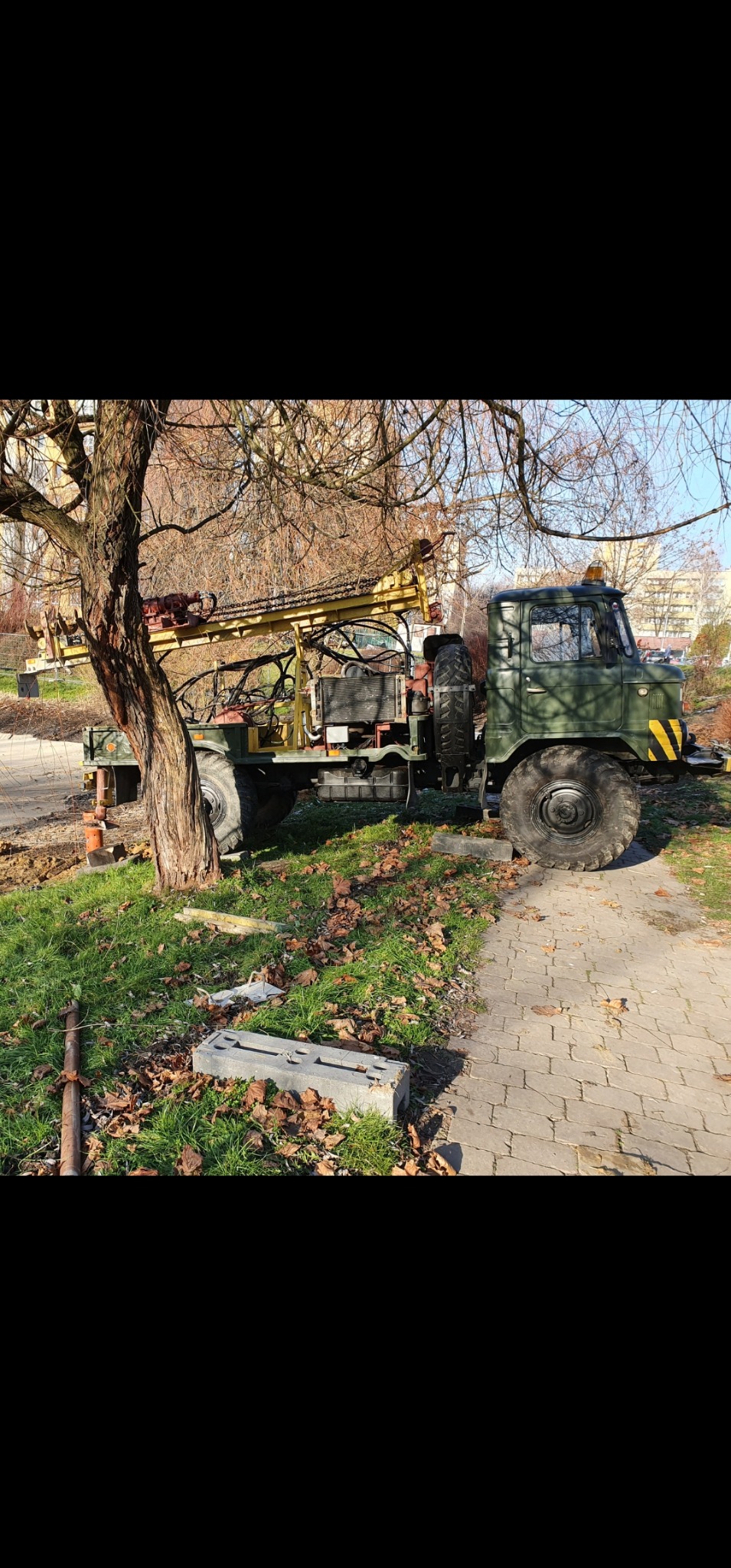 Zielona ciężarówka wiertnicza z żółtym masztem wiertniczym stoi obok drzewa i chodnika z kostki brukowej, w tle budynek mieszkalny.