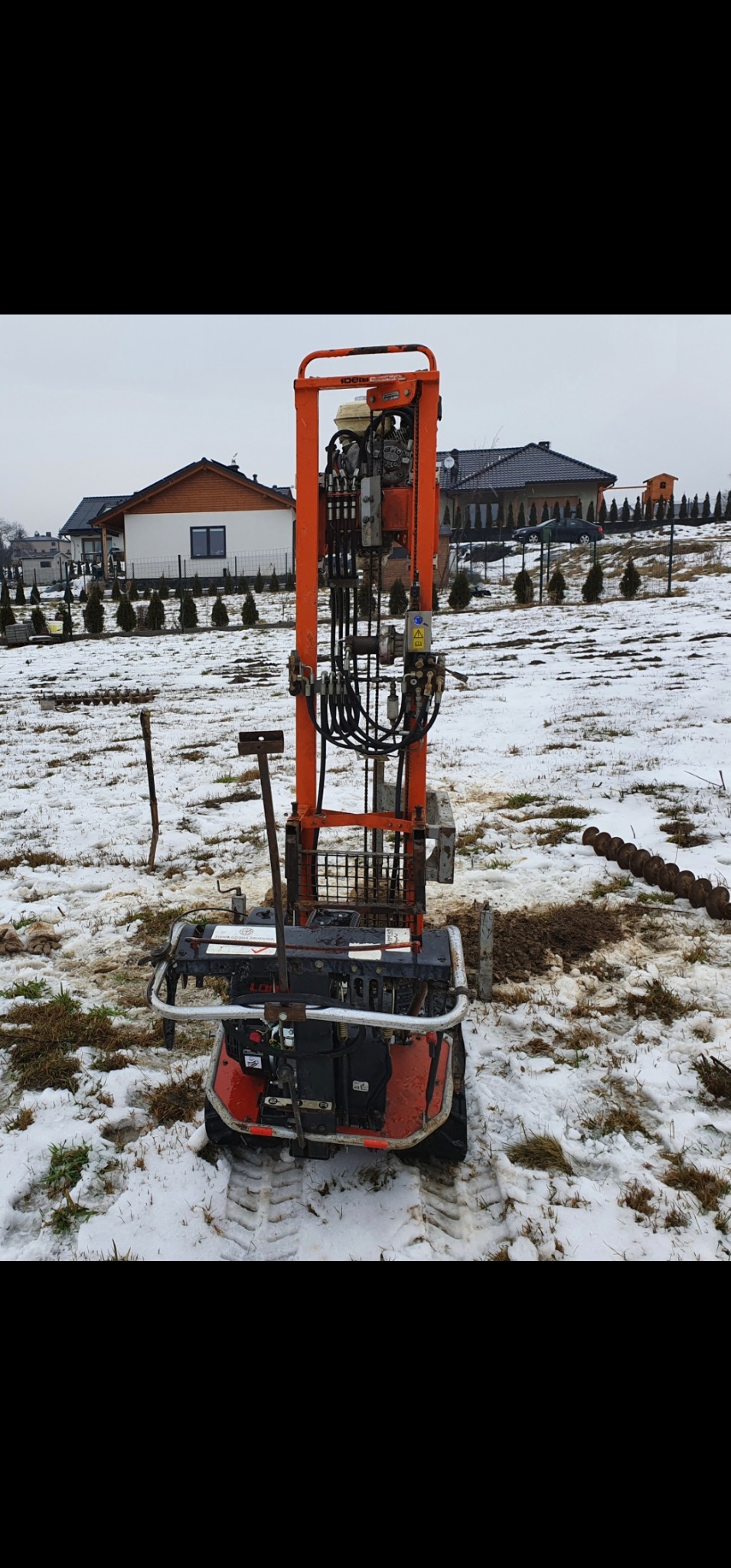 Wiertnica do studni na śniegu, widoczne ślady opon i elementy wiertła, w tle zabudowa jednorodzinna