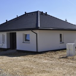 DOBRY DOM INVEST SPÓŁKA Z OGRANICZONĄ ODPOWIEDZIALNOŚCIĄ - Nowoczesny, parterowy dom z czarnym dachem i białymi ścianami, widoczny skrzynka elektryczna na zewnątrz, teren wokół w trakcie prac ziemnych.
