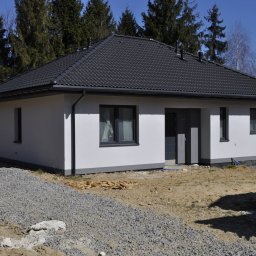 DOBRY DOM INVEST SPÓŁKA Z OGRANICZONĄ ODPOWIEDZIALNOŚCIĄ - Parterowy dom z ciemną dachówką i szarymi ramami okiennymi, otoczony ziemią i stertą żwiru przed wejściem.