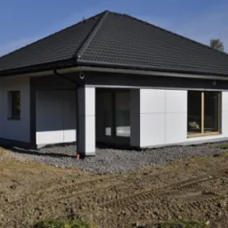 Nowoczesny parterowy dom z szarą dachówką, białymi panelami elewacyjnymi i dużymi oknami, teren wokół domu częściowo utwardzony żwirem, widoczne ślady opon na ziemi.