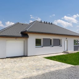 DOBRY DOM INVEST SPÓŁKA Z OGRANICZONĄ ODPOWIEDZIALNOŚCIĄ - Nowoczesny parterowy dom z garażem, jasna elewacja, ciemnoszara dachówka, kostka brukowa na podjeździe, trawnik, błękitne niebo z chmurami.