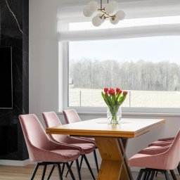 DOBRY DOM INVEST SPÓŁKA Z OGRANICZONĄ ODPOWIEDZIALNOŚCIĄ - Jasna jadalnia z drewnianym stołem na metalowej podstawie, otoczonym różowymi krzesłami z czarnymi nogami, z widokiem na krajobraz za oknem i nowoczesną lampą sufitową.
