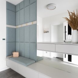 DOBRY DOM INVEST SPÓŁKA Z OGRANICZONĄ ODPOWIEDZIALNOŚCIĄ - Wnęka w przedpokoju z siedziskiem obitym jasnoniebieską tkaniną, panelami ściennymi w tym samym kolorze i złotymi haczykami; na siedzisku leży mała, beżowa torebka.