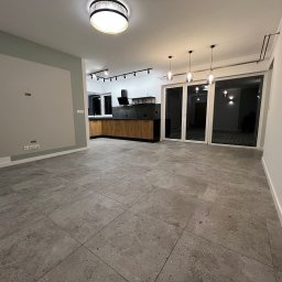 DOBRY DOM INVEST SPÓŁKA Z OGRANICZONĄ ODPOWIEDZIALNOŚCIĄ - Nowoczesne wnętrze domu z szarą podłogą, drewnianą zabudową kuchenną i dużymi przesuwnymi drzwiami tarasowymi, oświetlone punktowymi lampami sufitowymi.