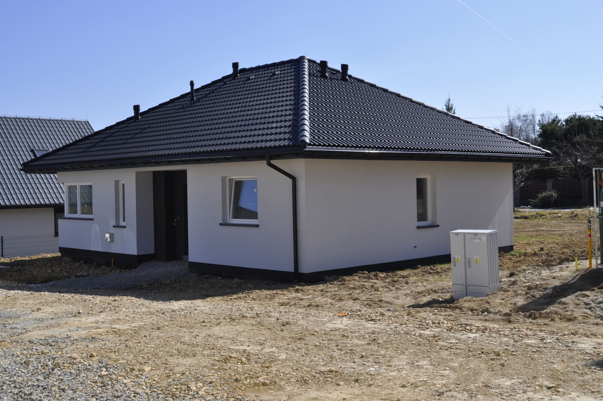 Nowoczesny, parterowy dom z czarnym dachem i białymi ścianami, widoczny skrzynka elektryczna na zewnątrz, teren wokół w trakcie prac ziemnych.