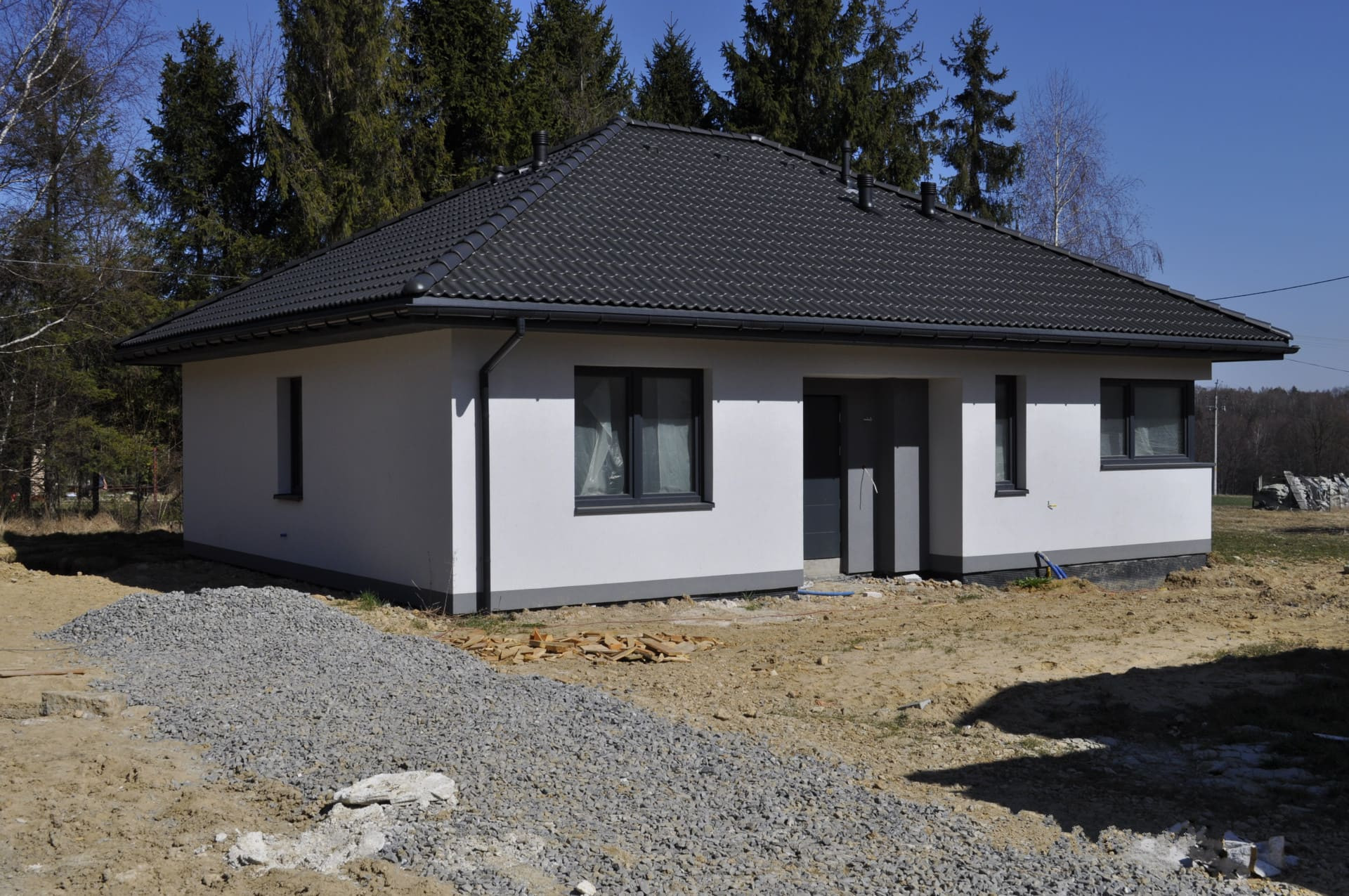 Parterowy dom z ciemną dachówką i szarymi ramami okiennymi, otoczony ziemią i stertą żwiru przed wejściem.