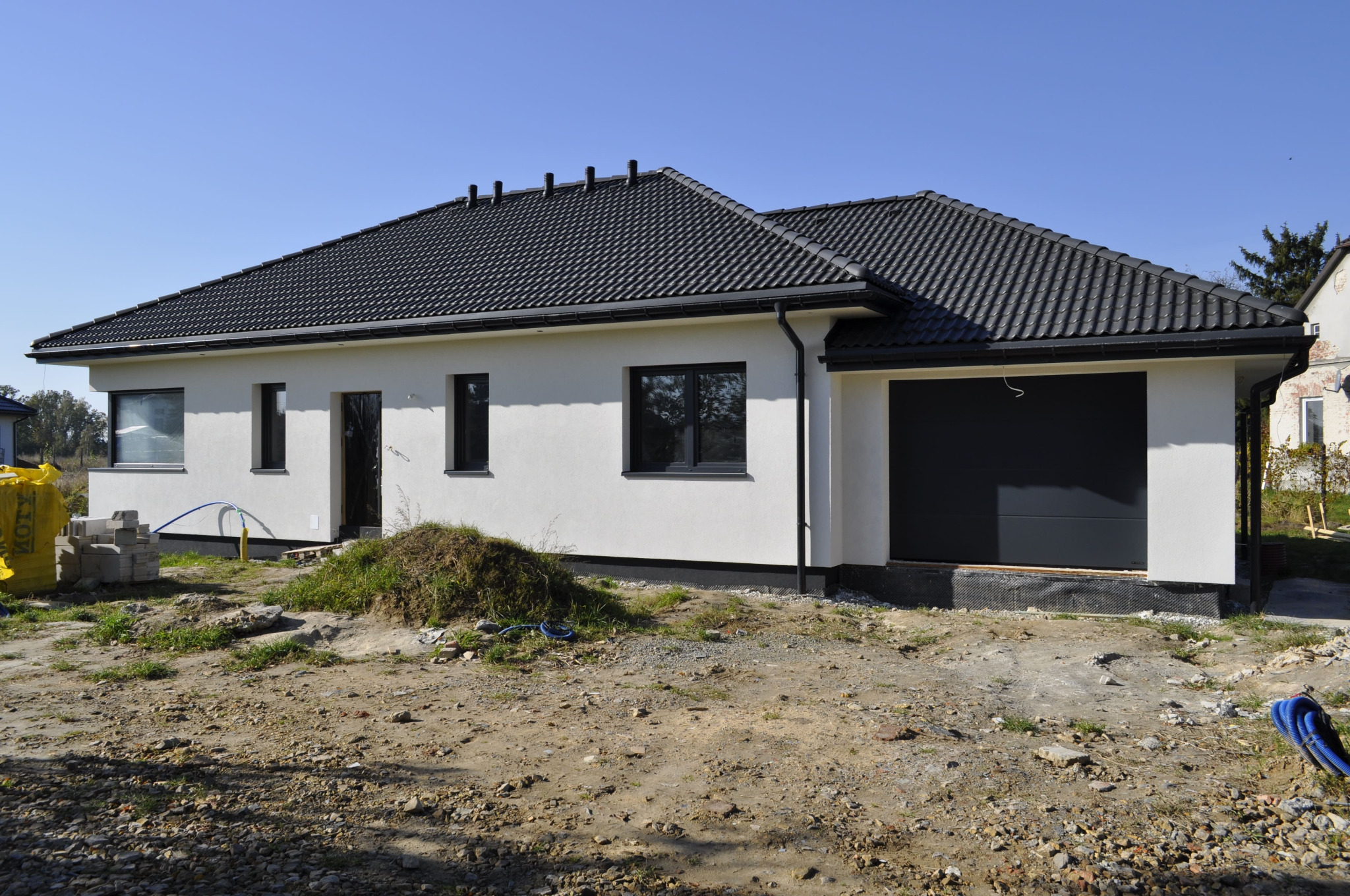 Parterowy dom z ciemną dachówką i garażem w trakcie wykończenia, widoczne materiały budowlane i niezagospodarowany teren wokół.