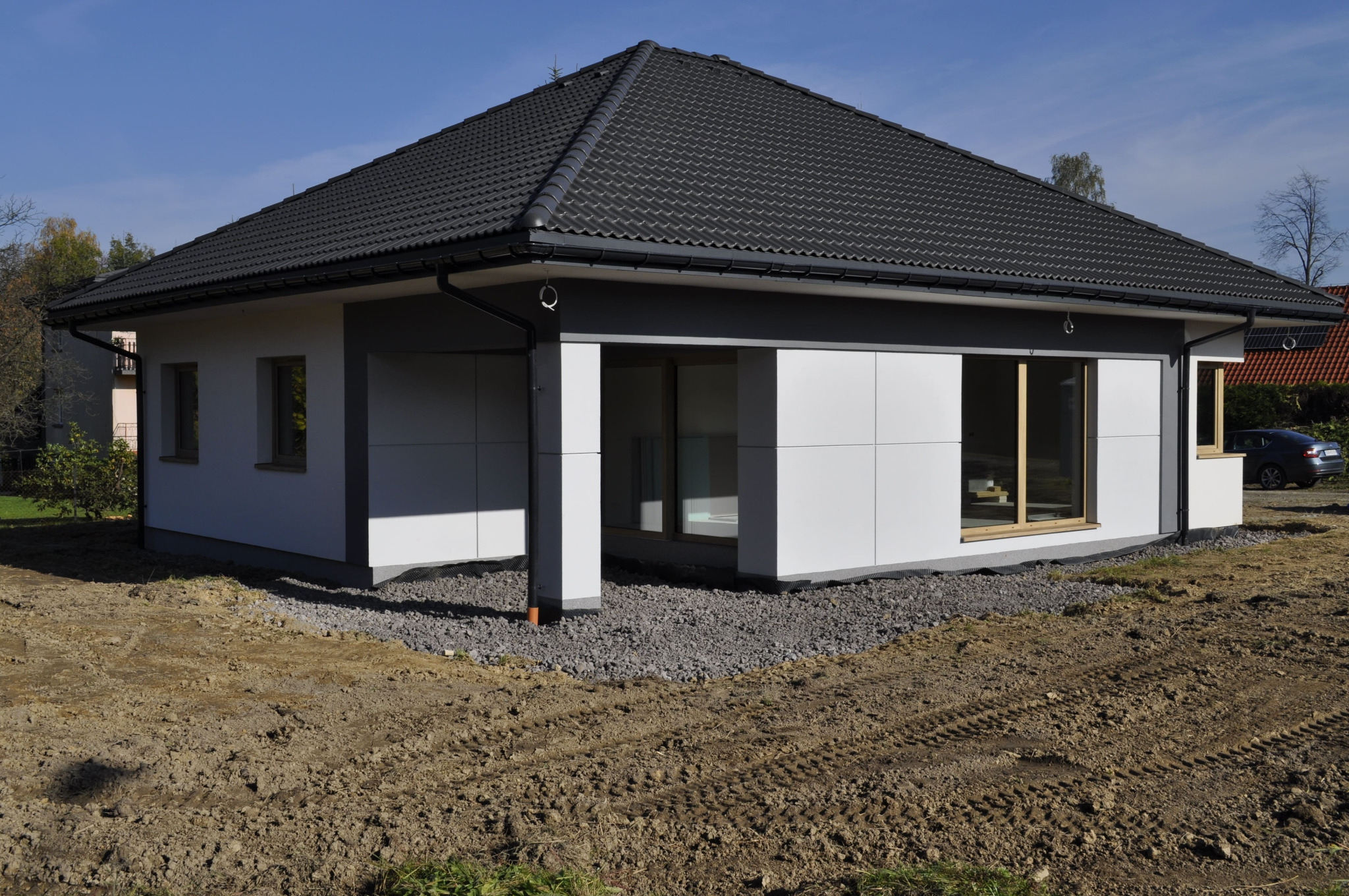 Nowoczesny parterowy dom z szarą dachówką, białymi panelami elewacyjnymi i dużymi oknami, teren wokół domu częściowo utwardzony żwirem, widoczne ślady opon na ziemi.