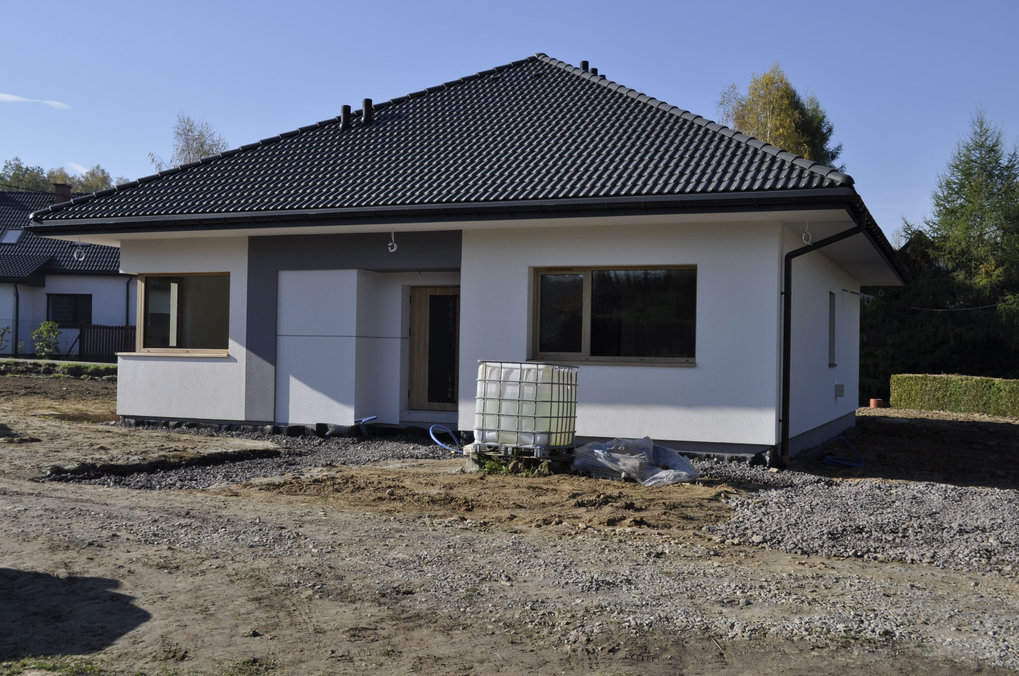 Niedokończony parterowy dom z ciemną dachówką, jasną elewacją i drewnianymi oknami, z zbiornikiem na wodę przed wejściem i przygotowanym terenem pod ogród.