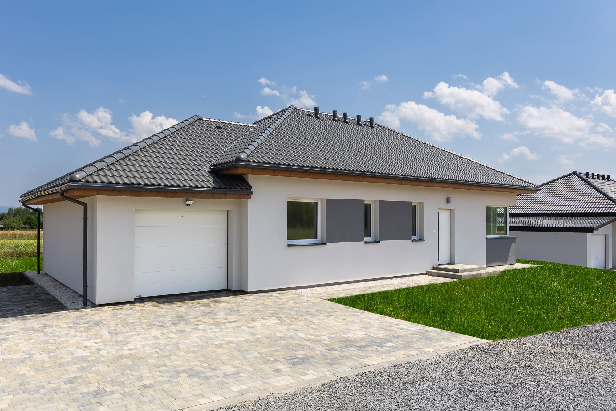 Nowoczesny parterowy dom z garażem, jasna elewacja, ciemnoszara dachówka, kostka brukowa na podjeździe, trawnik, błękitne niebo z chmurami.